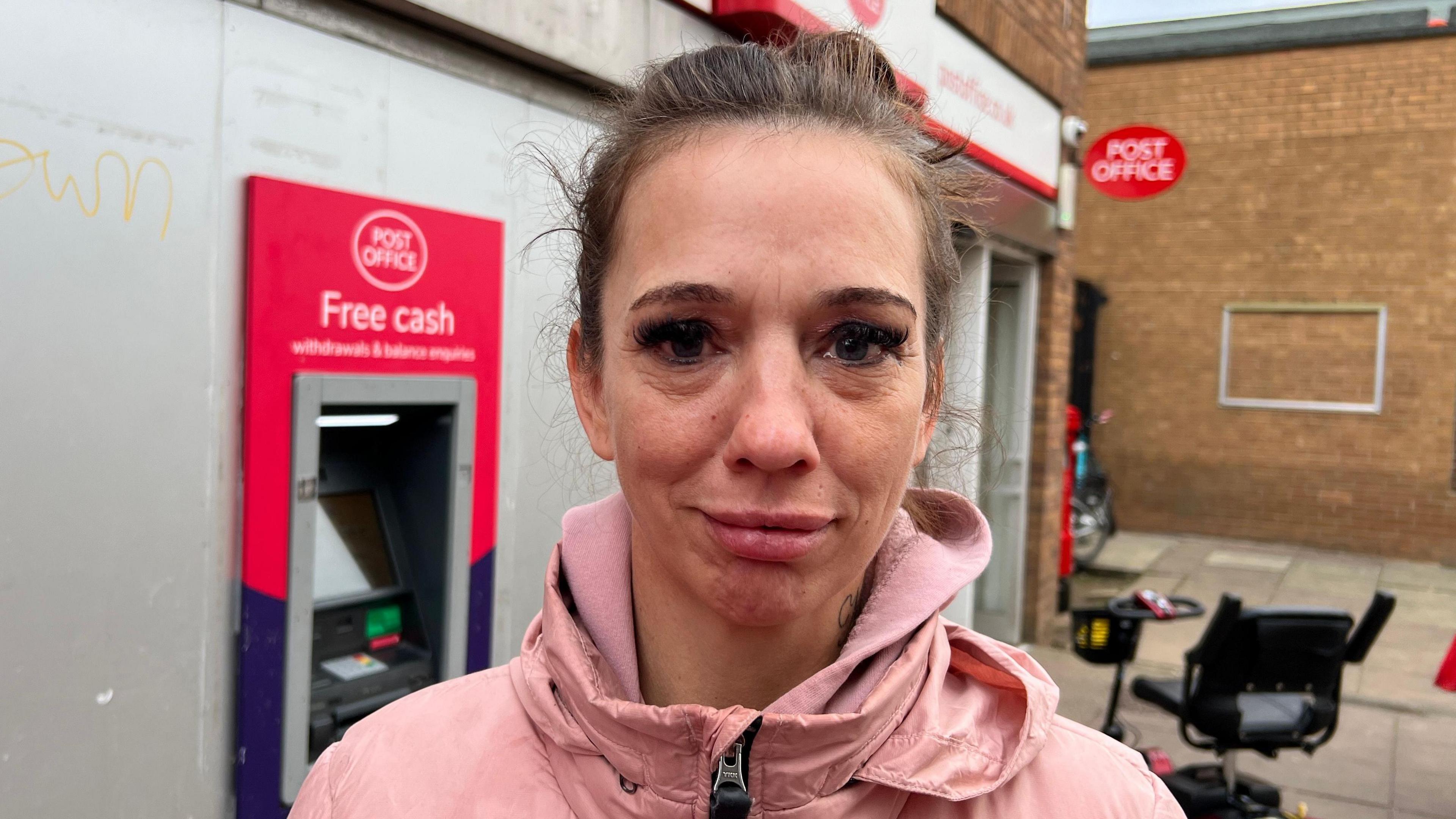 A woman with dark brown hair pulled back with a hair band, wearing a pink zip up coat. She is standing outside Bransholme Post Office and there is a cash machine and mobility scooter in the background.