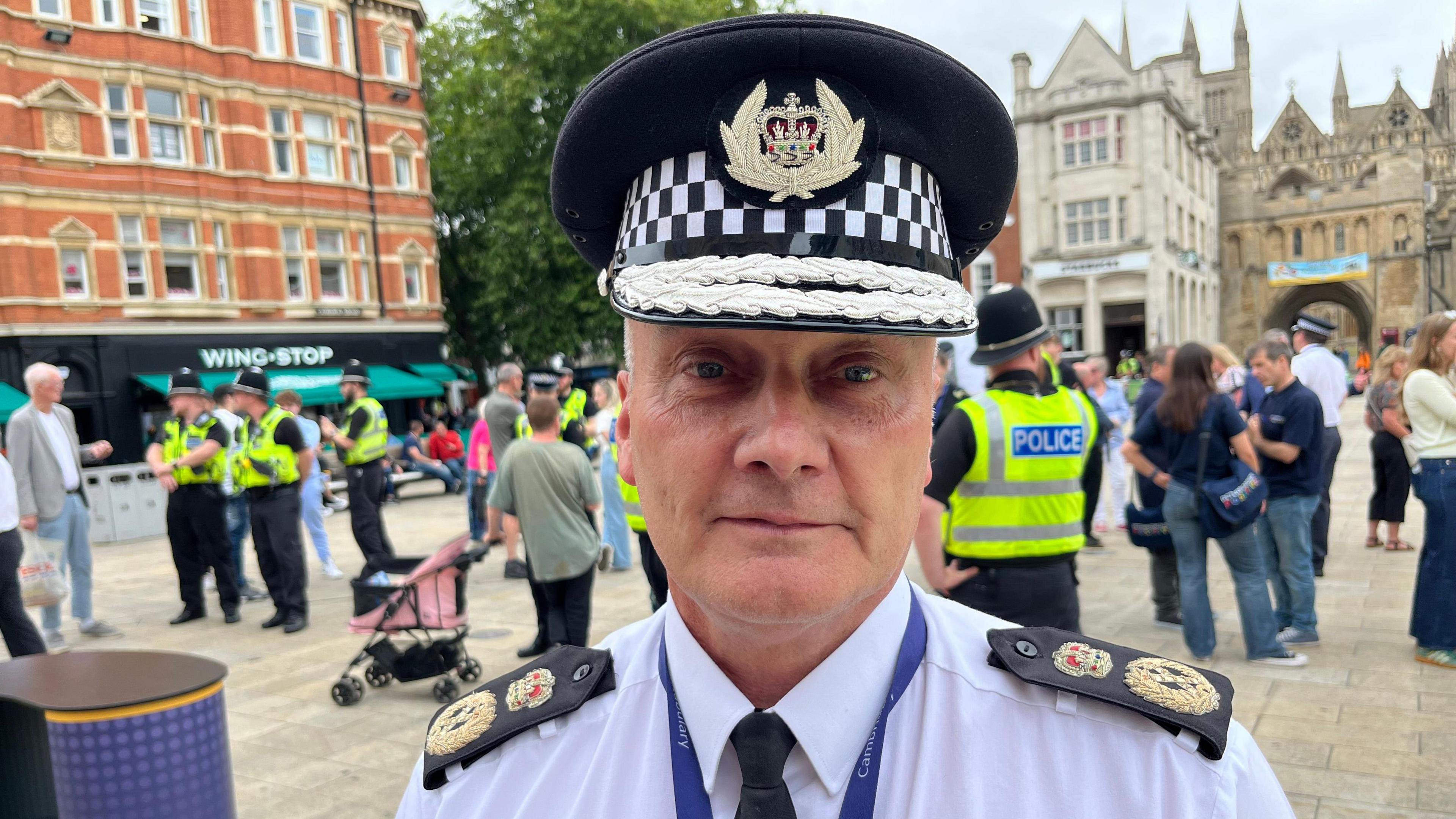 Chief Constable Nick Dean stands in Peterborough square with officers and the general public behind him. Trees and shops can be seen in the distance. The chief constable stares at the camera. He wears a white shirt with lapels denoting the Cambridgeshire force and his rank. He also has on a black tie and a lanyard round his neck.