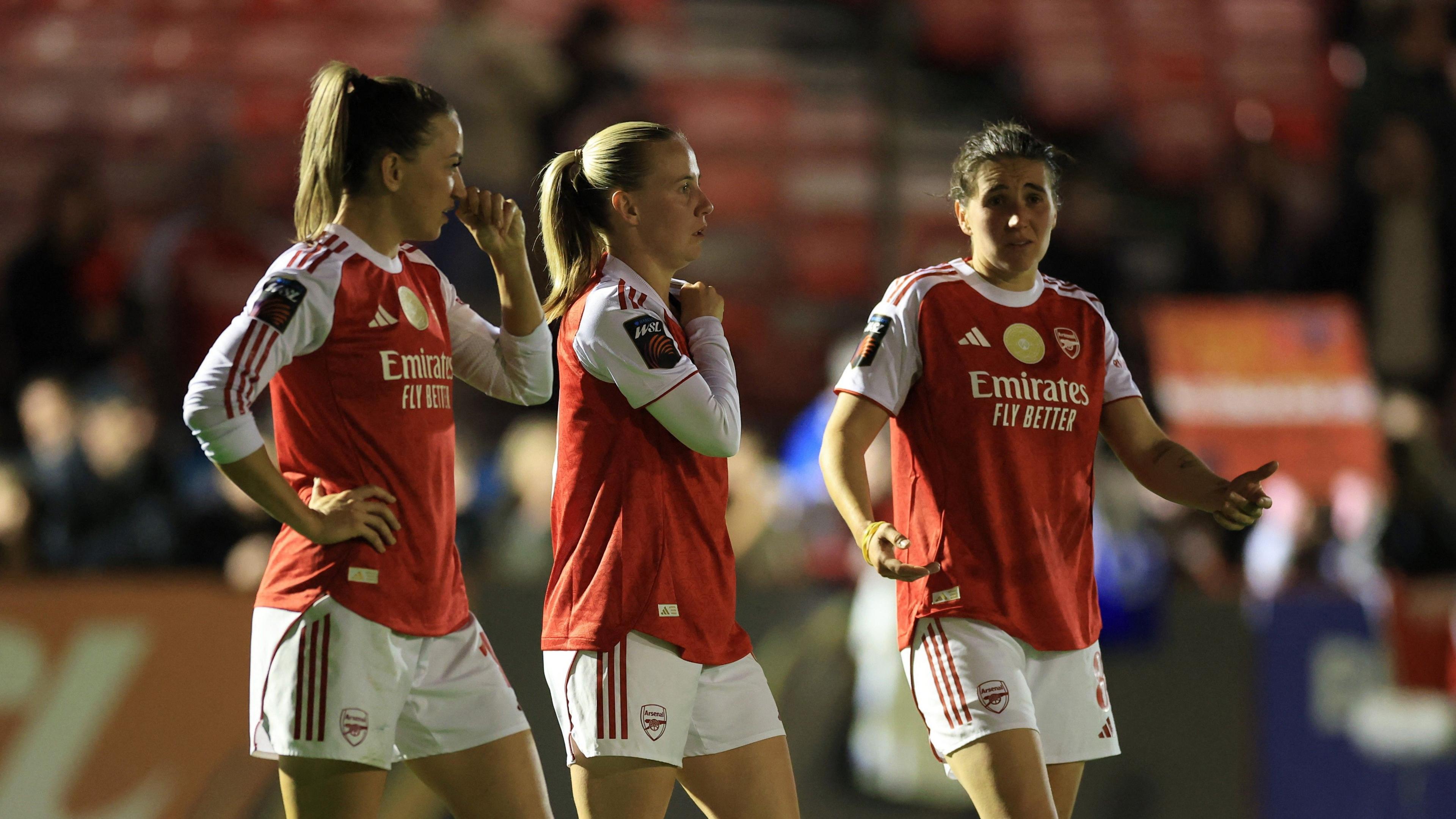 Three Arsenal players, wearing red and white shirts and white shorts, look dejected after the final whistle