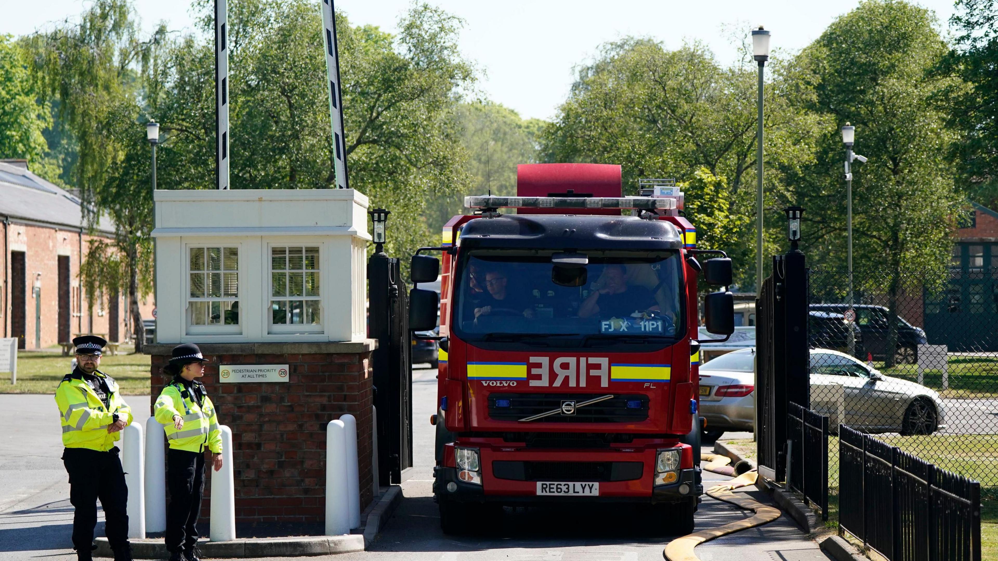 Fire engine driving out of barriers with two police officers in high-vis standing to the left. There are trees, buildings and vehicles in the background