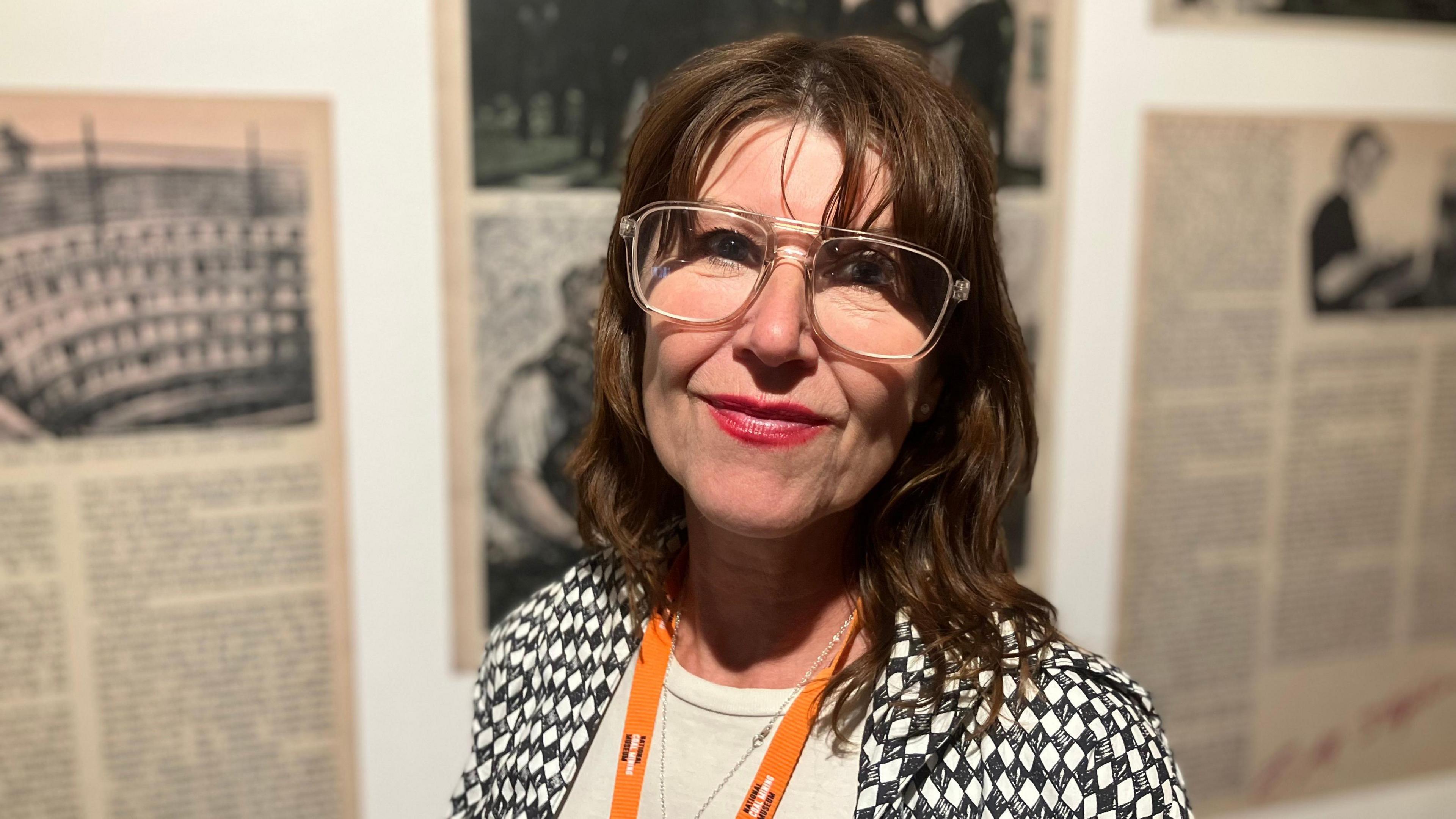 A woman with dark hair and glasses smiles at the camera. She wears a checked shirt with an orange lanyard. Behind her, newspaper articles hung on the wall are blurred.