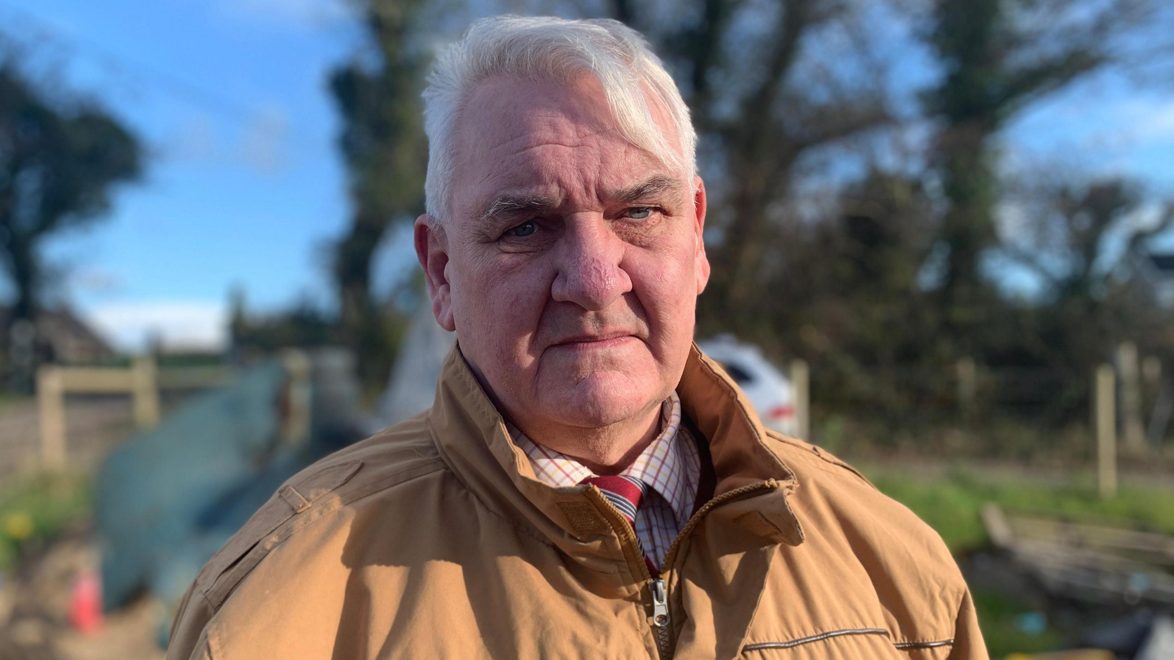 A man with short grey hair is looking at the camera. He is wearing a pale brown coat and has a check shirt and red tie on underneath.