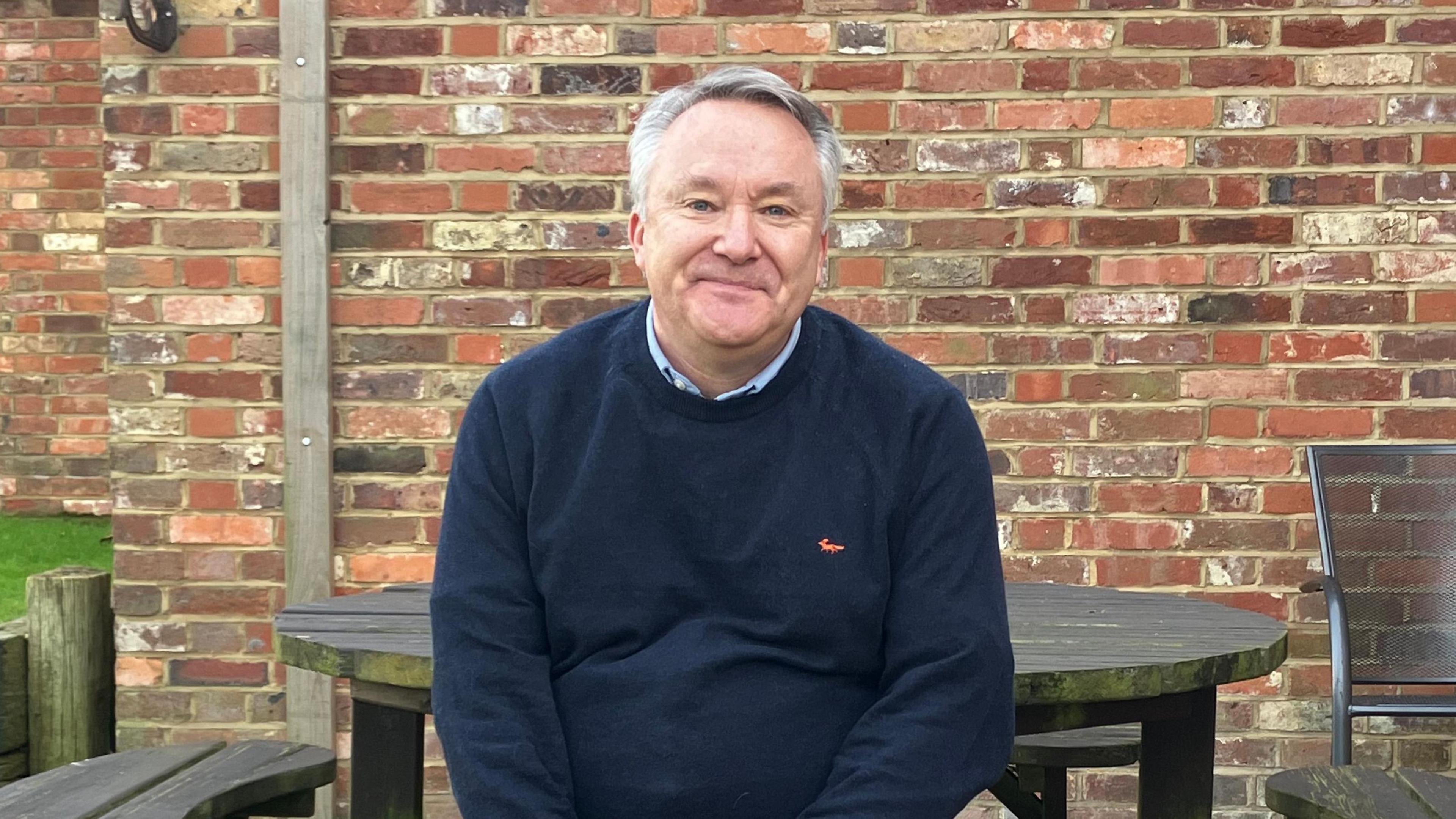 Stuart Wilson is sat down at a picnic table and has grey/white hair. He is sat in a pub beer garden and is wearing a dark blue jumper with light brown trousers. Behind him is a brick wall. 