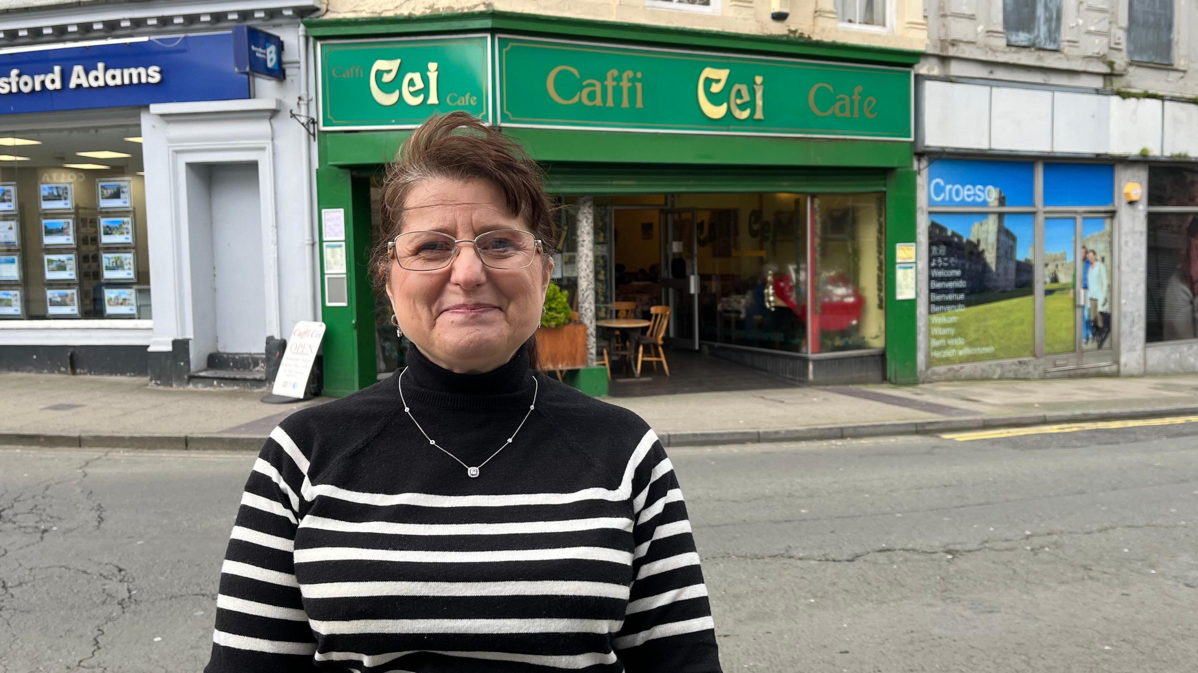 Gaynor Morris standing opposite Caffi Cei which has a green store front sign and writing in gold-coloured letters. She is wearing a black and white striped jumper and a silver necklace and has brown hair tied back.