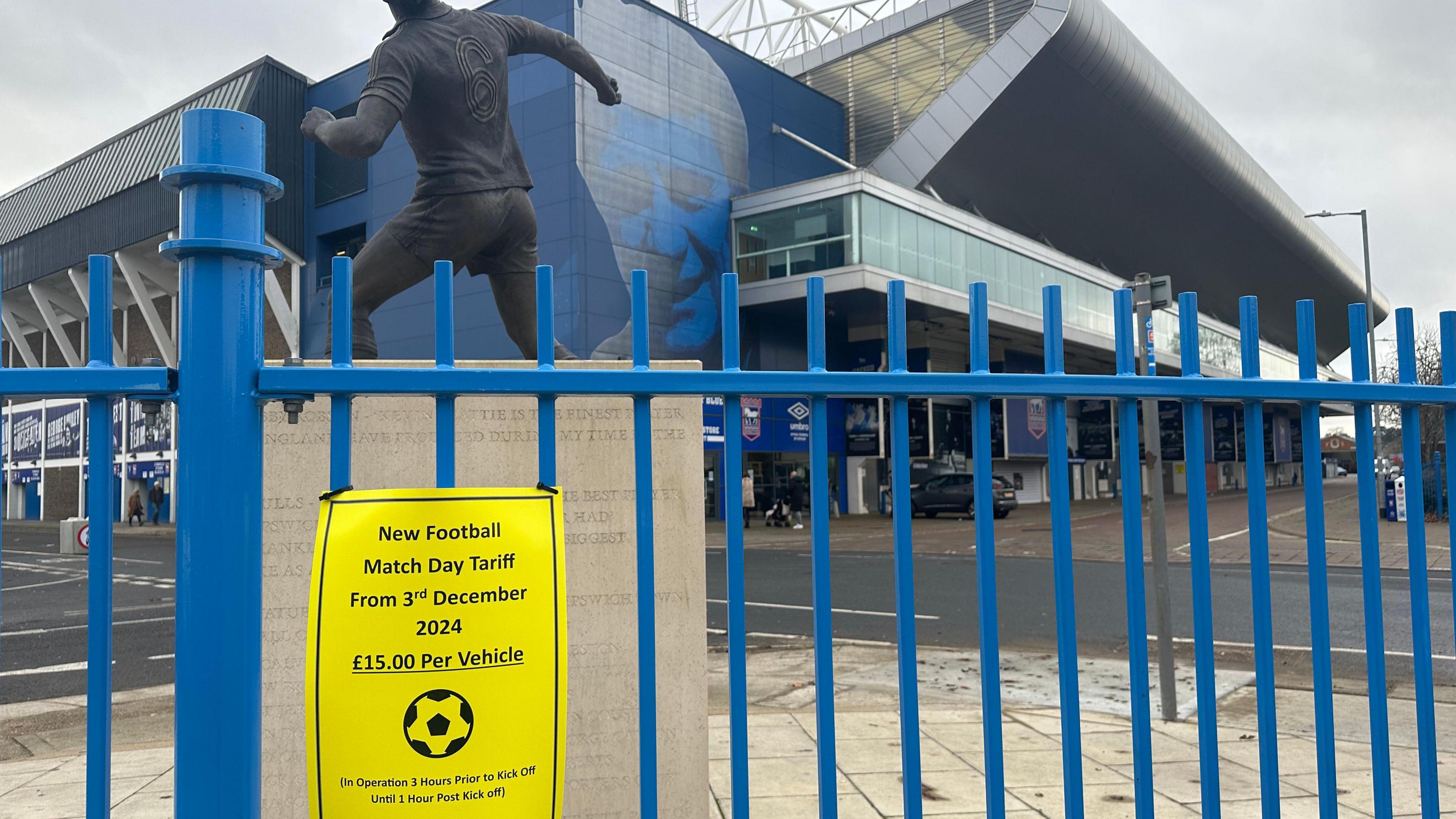 The new parking tariff sign sits on a blue fence. Behind the fence is a statue of a football player with the stadium sat further behind that. A road around the stadium can also been seen.