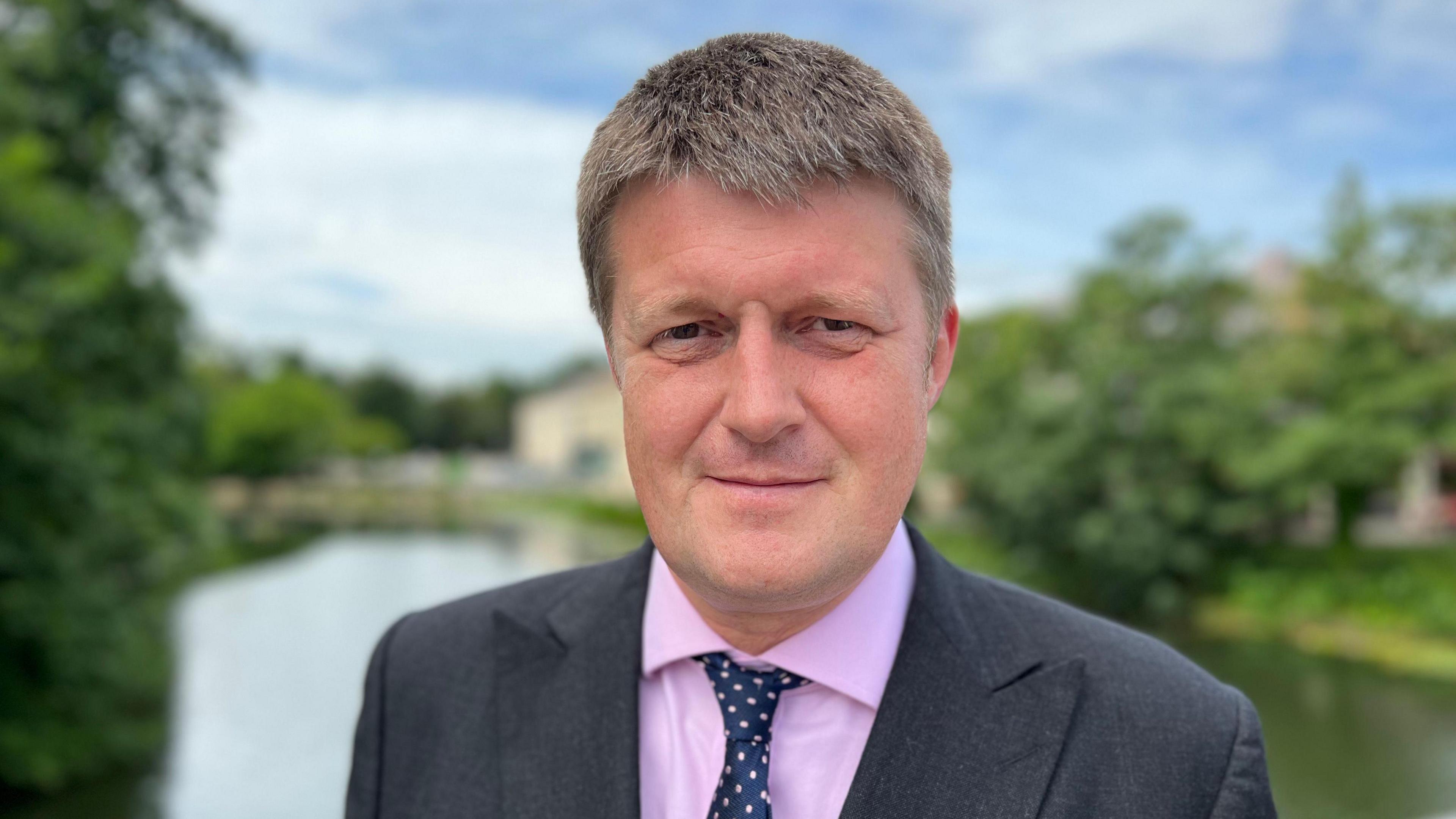 Richard Clewer in a pink shirt and dark suit standing in front of a body of water (not the pothole)