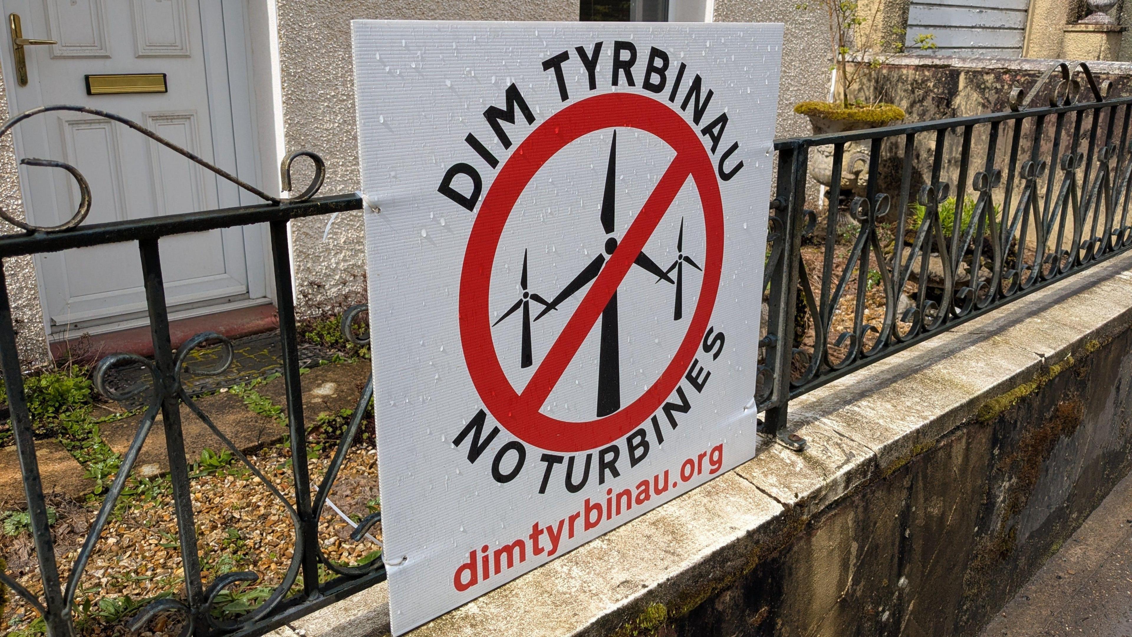 Poster ar glwyd o flaen  tŷ ym mhentref Rhydcymerau sy'n dweud 'Dim Tyrbinau'. Mae'r poster yn dangos tyrbinau ac arwydd coch drostyn nhw, yn cefnogi eu gwahardd.