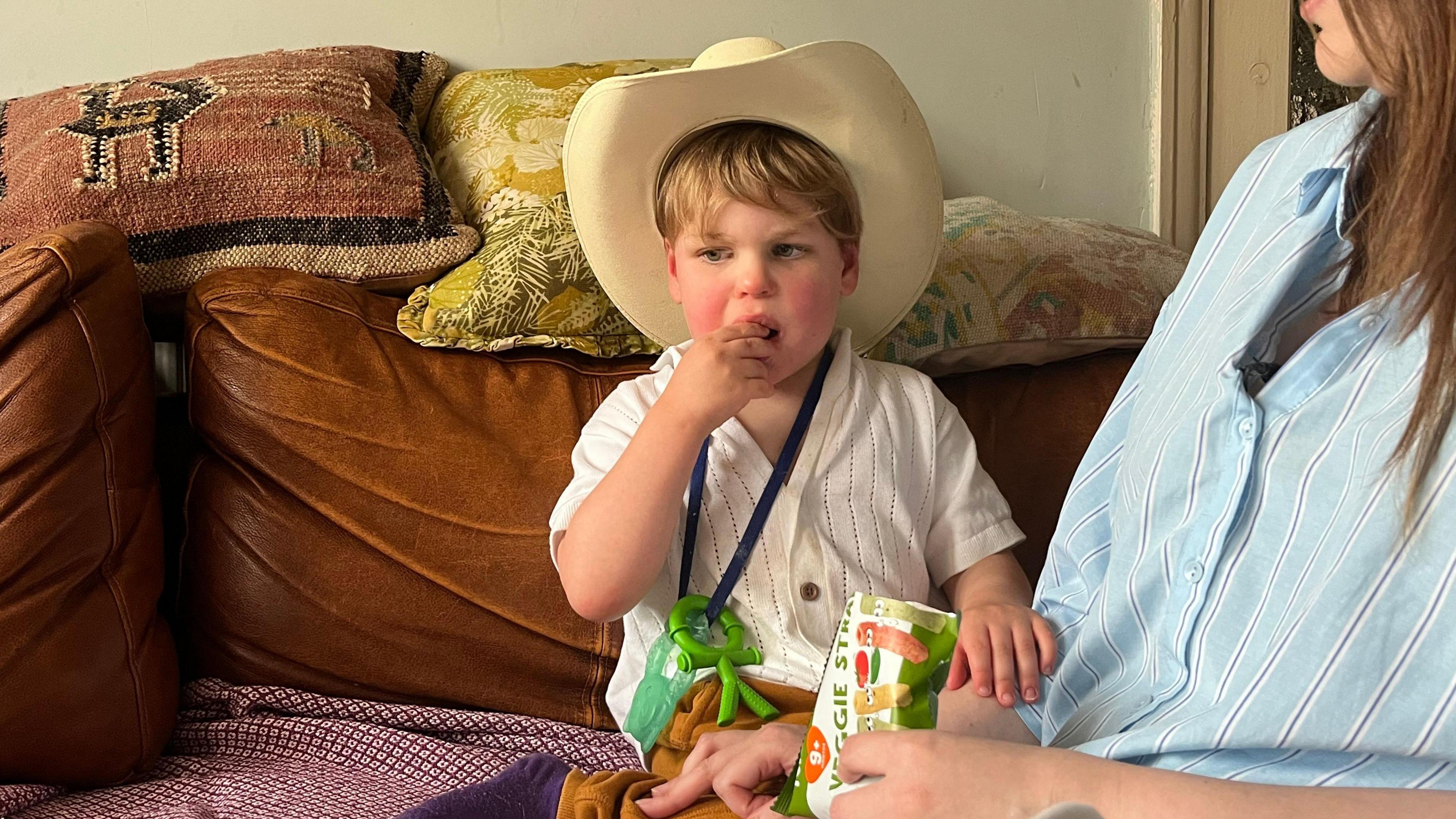 Ivan wearing a cowboy hat and eating a snack. He has a white shirt on and a blue lanyard around his neck with a green plastic shape on it. A woman with brown hair can just be seen in the foreground, holding a packet of snacks.