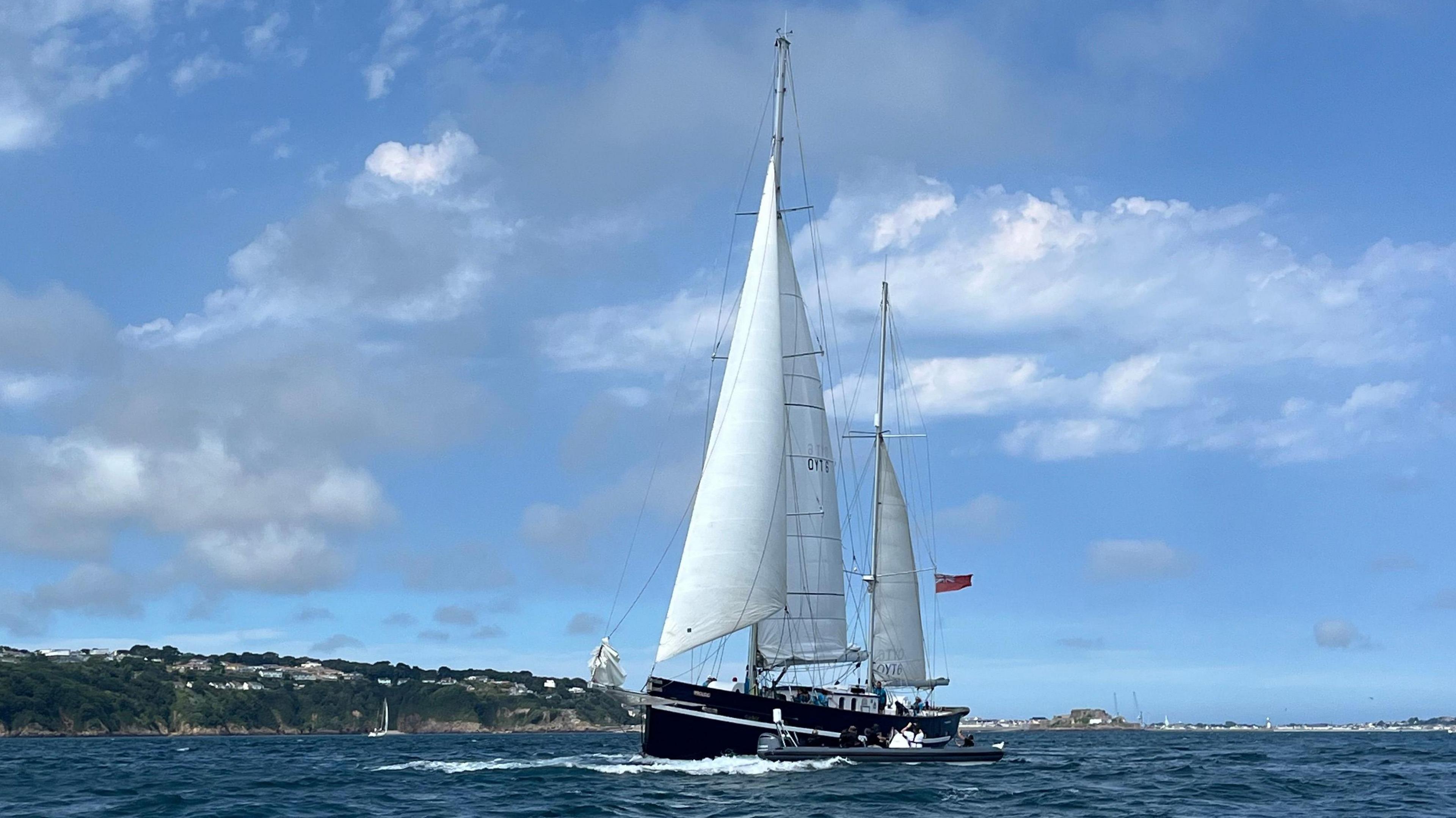 A large sailing boat with white sails in the sea with the coast of Guernsey in the background, blue skies with grey and white clouds