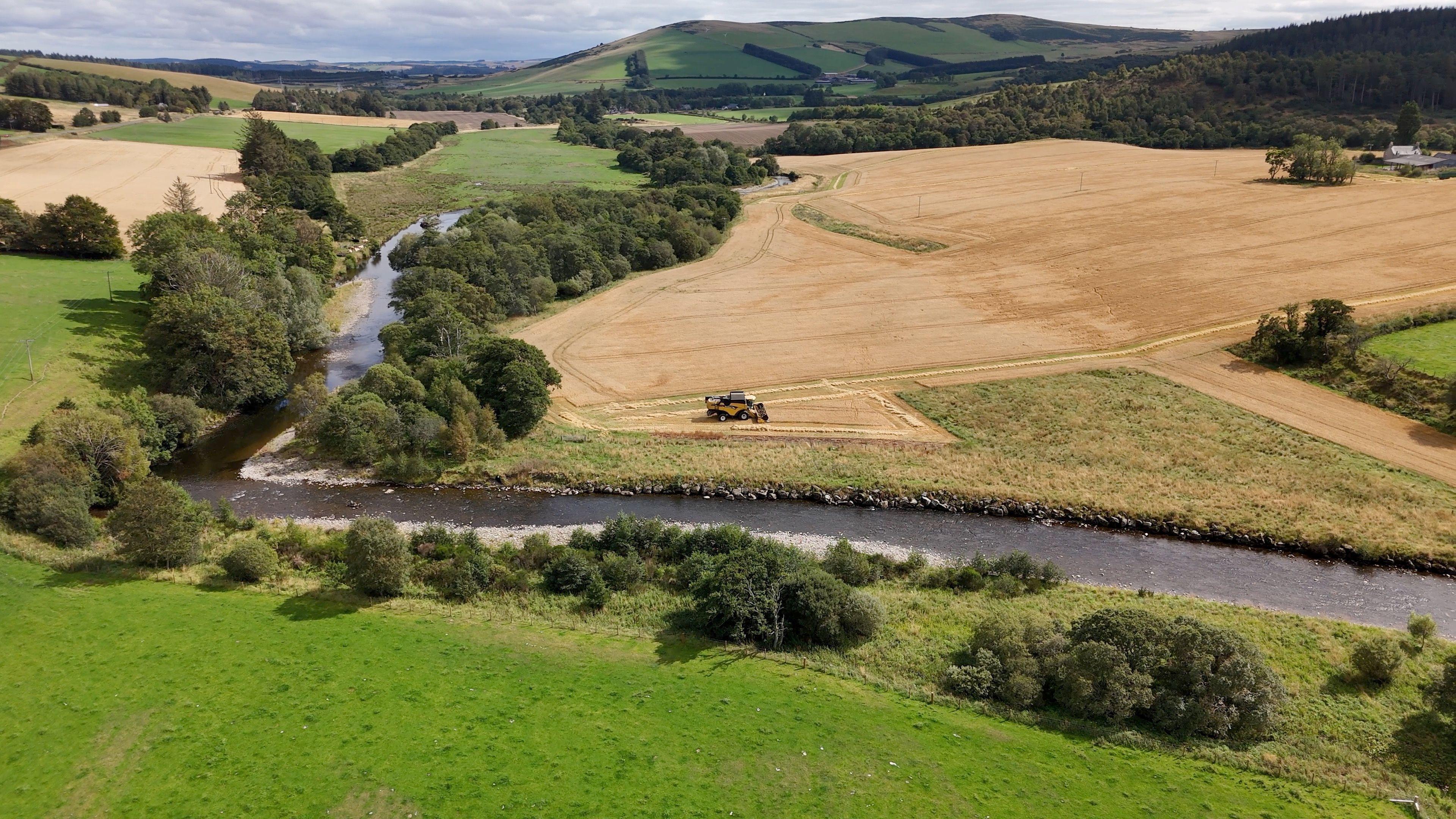 An aerial shot of the Deveron, with parts of the river bed showing. There is a golden field at the top right of the picture with a farm vehicle on it, next to some dry land, and there is a green field at the bottom