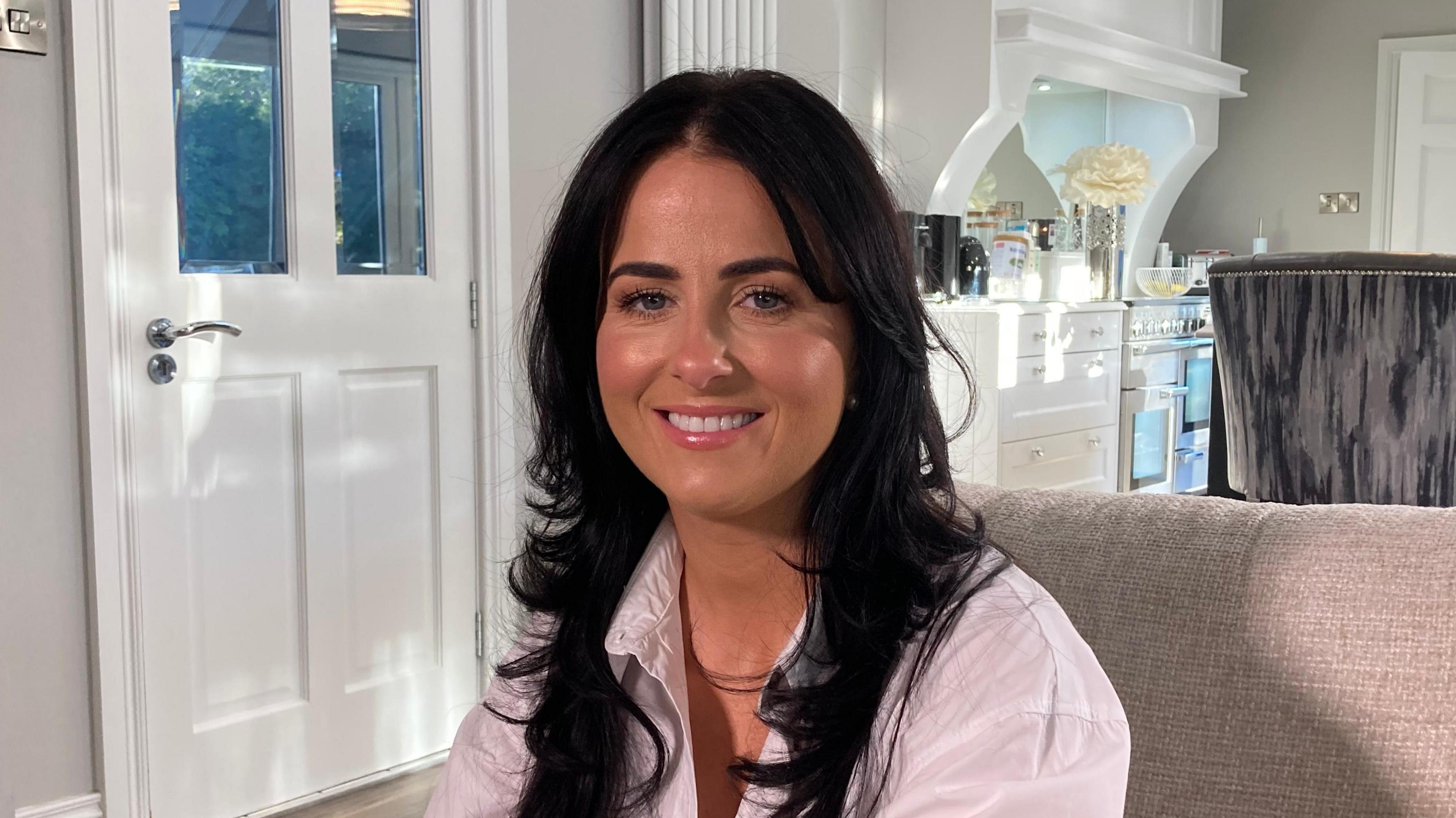 Lisa McHugh has long, black, wavy hair in a front parting. She is looking directly into the camera and smiling. She is wearing a white shirt and is sitting on a grey sofa. A light-coloured kitchen is visible in the background. There is a white door to the left of the picture.