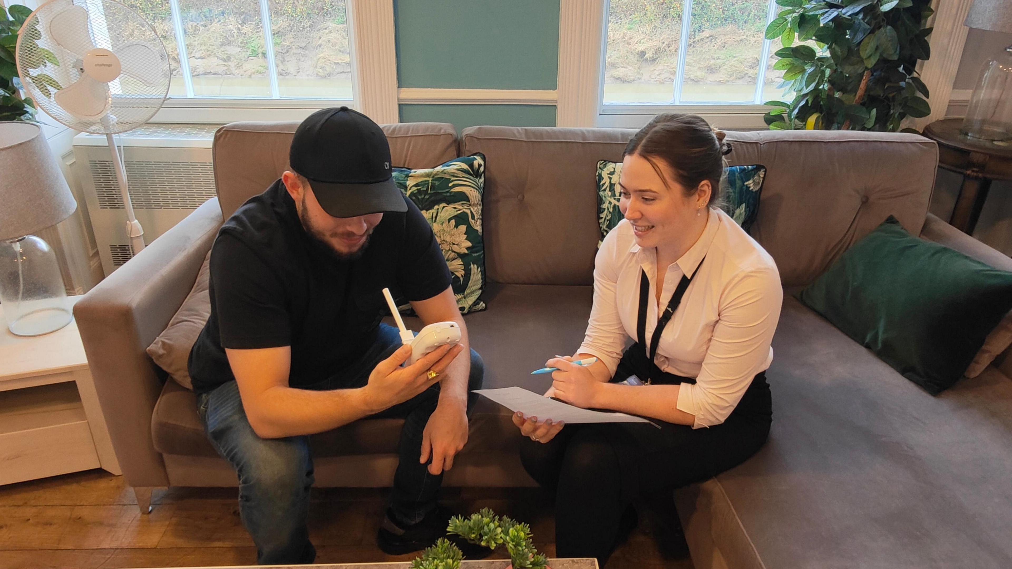 A man and a woman are sitting on a sofa, the man has a handheld breath monitor in his right hand, the woman is holding a pen and paper.