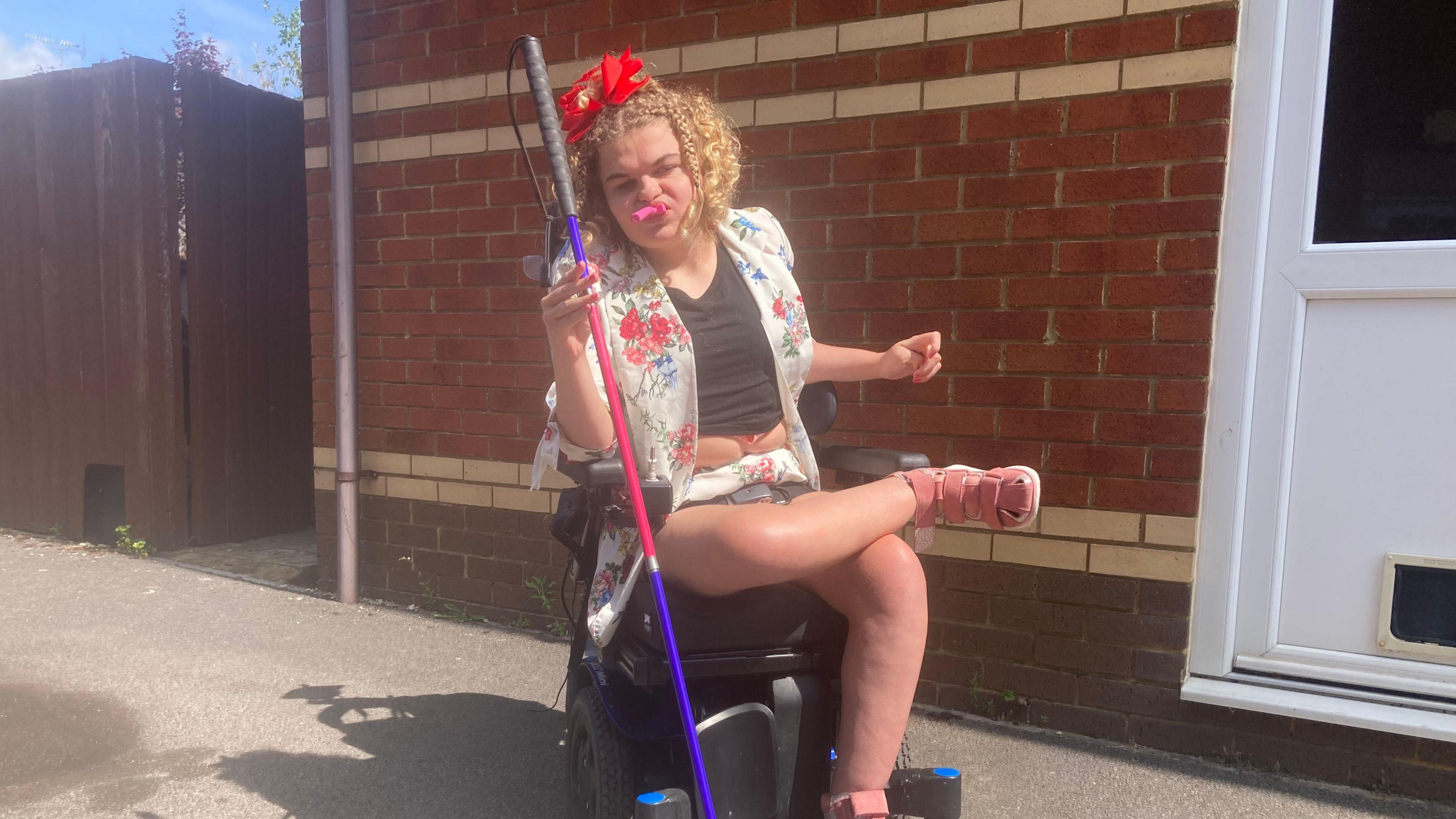 A woman sitting in a wheelchair holding a mobility stick in her right hand and a pink party blower in her mouth.