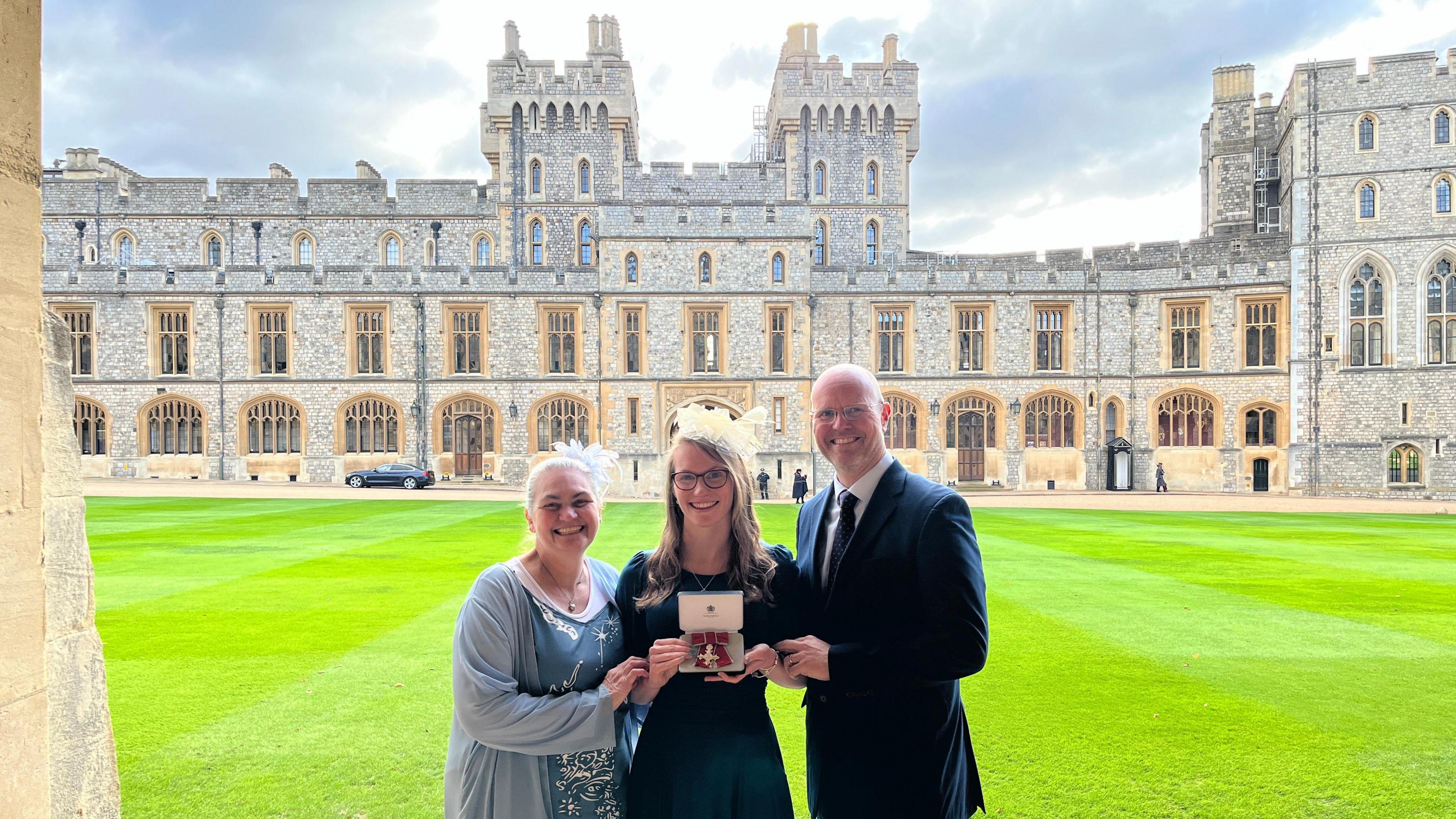 A woman on the left is wearing a blue chiffon dress with silver beading. In the middle is Rebecca, who is wearing glasses and a dark blue short sleeve dress holding a box containing her MBE. Standing on her right a tall bald man is in a dark suit and tie. Behind them are the grounds of Windsor Castle. 