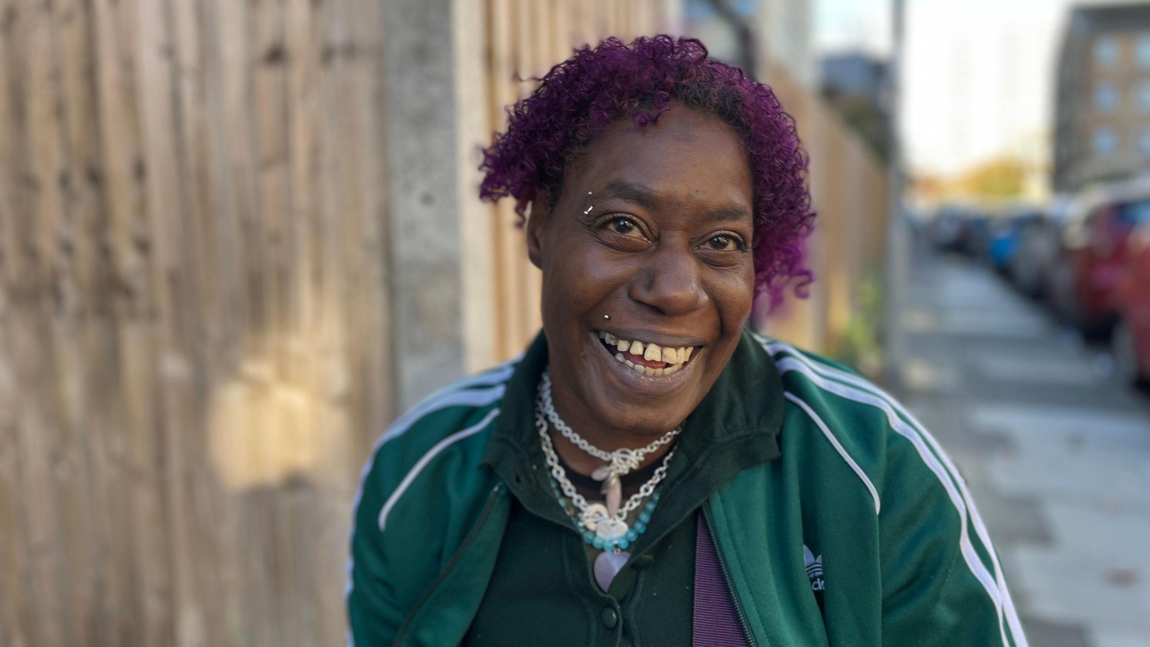 Kimberley Roberts wearing a dark green Adidas tracksuit. She has purple curly hair and several necklaces on her neck. She has a lip and eyebrow piercing and is smiling at the camera