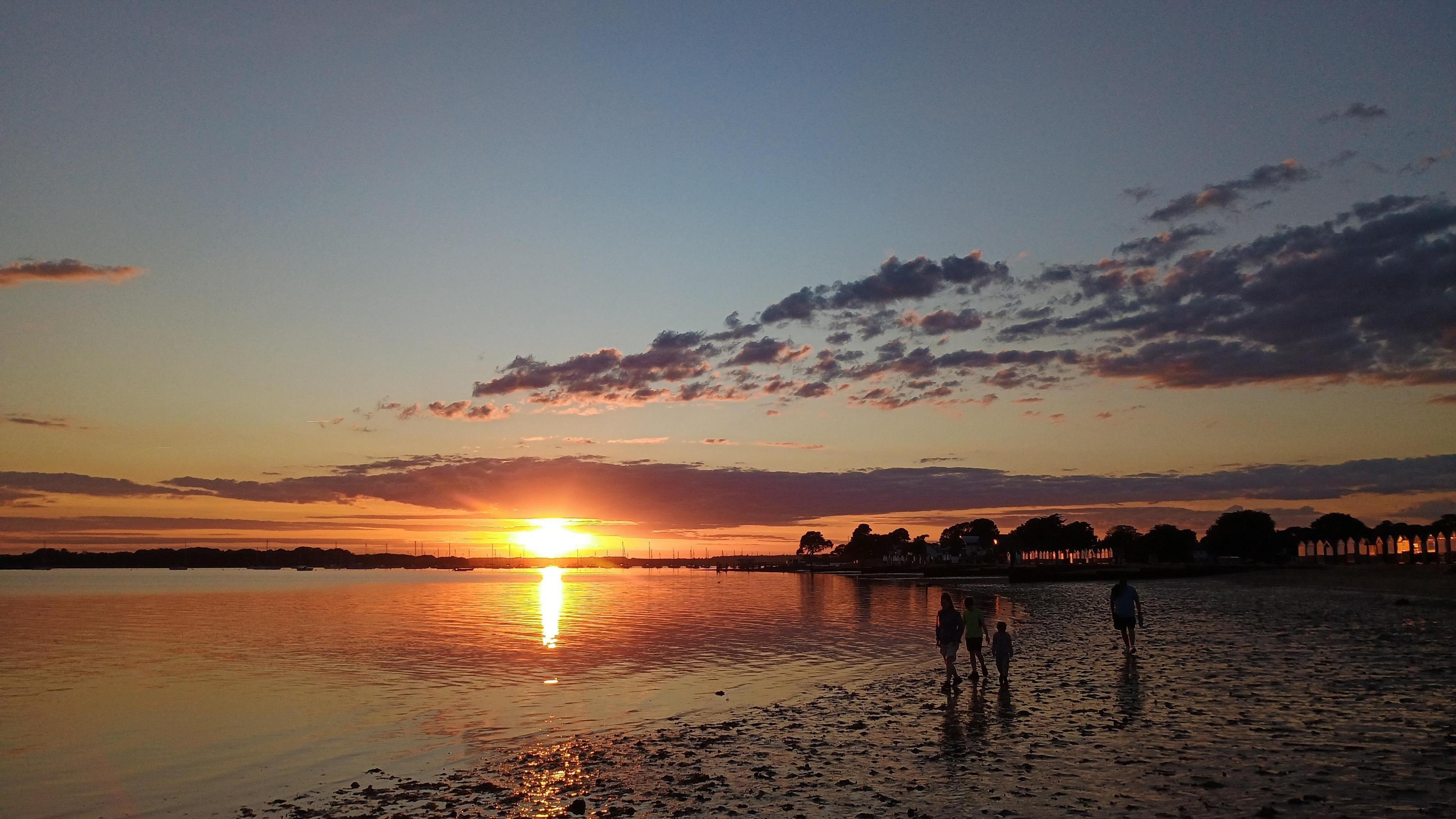 The golden sun sets in the background over a low tide and beach. A tree line can be seen in the background. A family is seen in silhouette walking along the sea's edge across the mudflats.