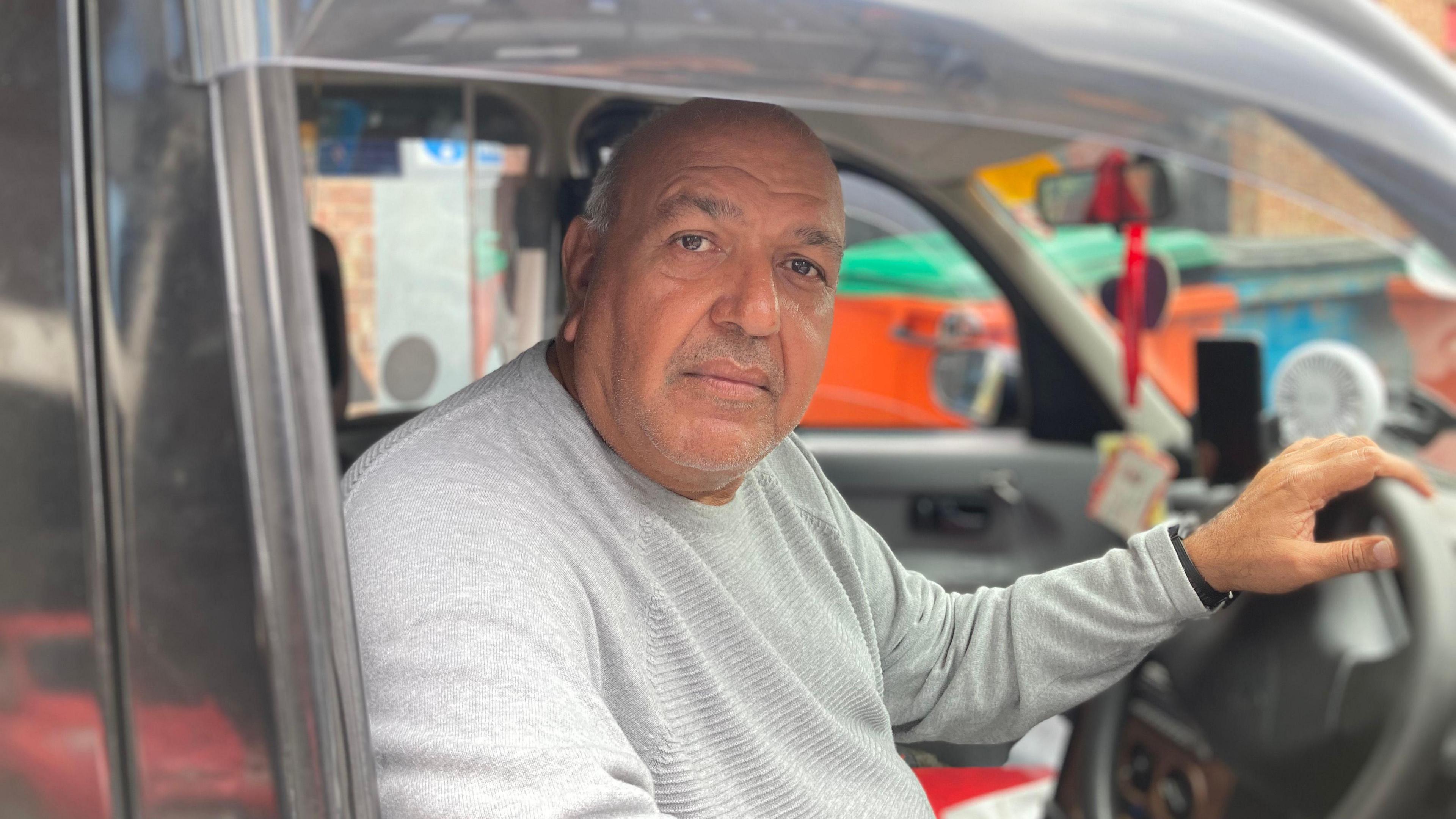 A man looking through the driver's window of his taxi in Coventry. He has a hand on the steering wheel.
