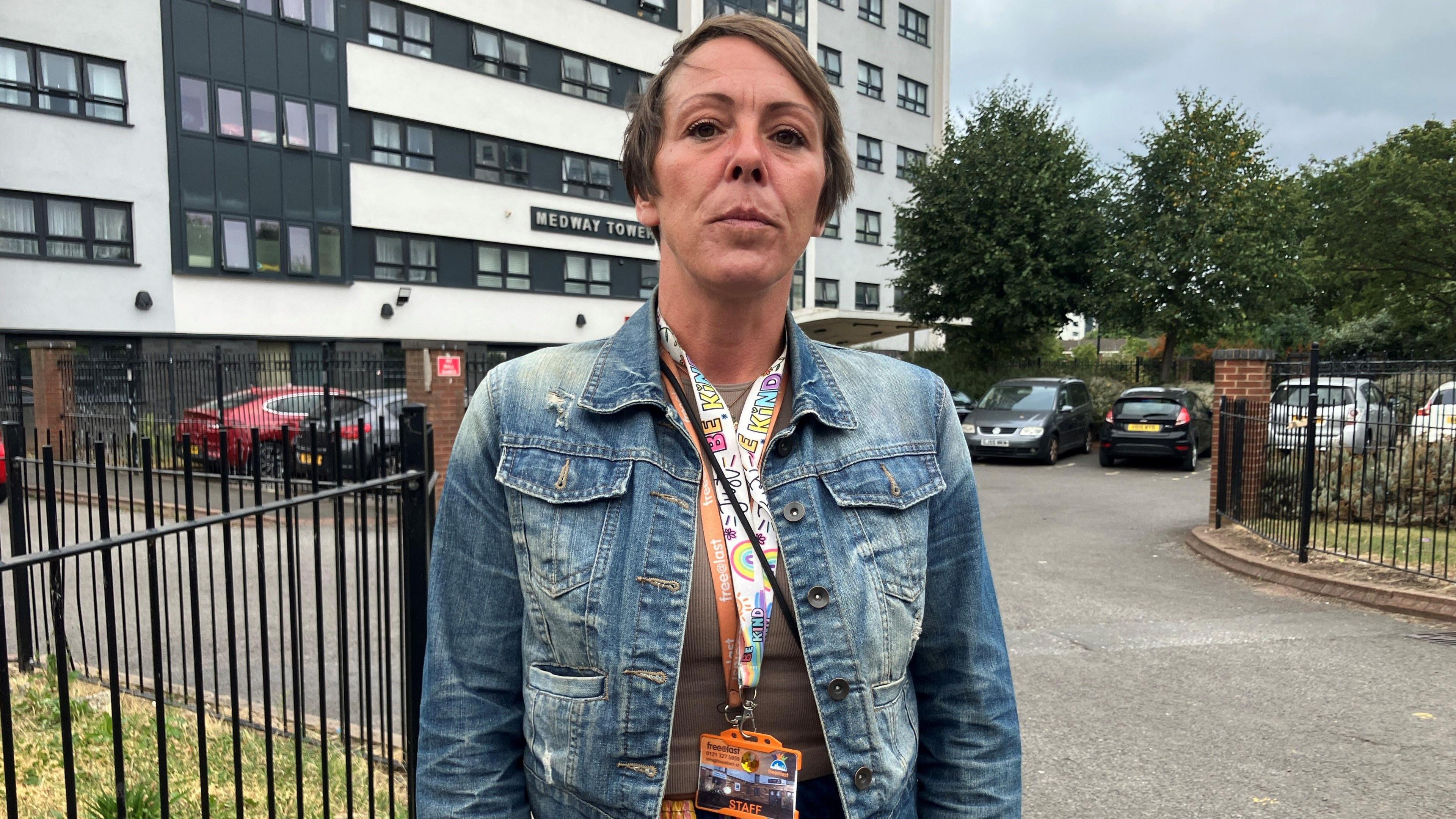 Woman looking angrily at camera. She has short brown hair, and is wearing a denim jacket and coloured lanyards. She is standing in front of a black and white tower block, with a sign reading Medway Tower on the front.  