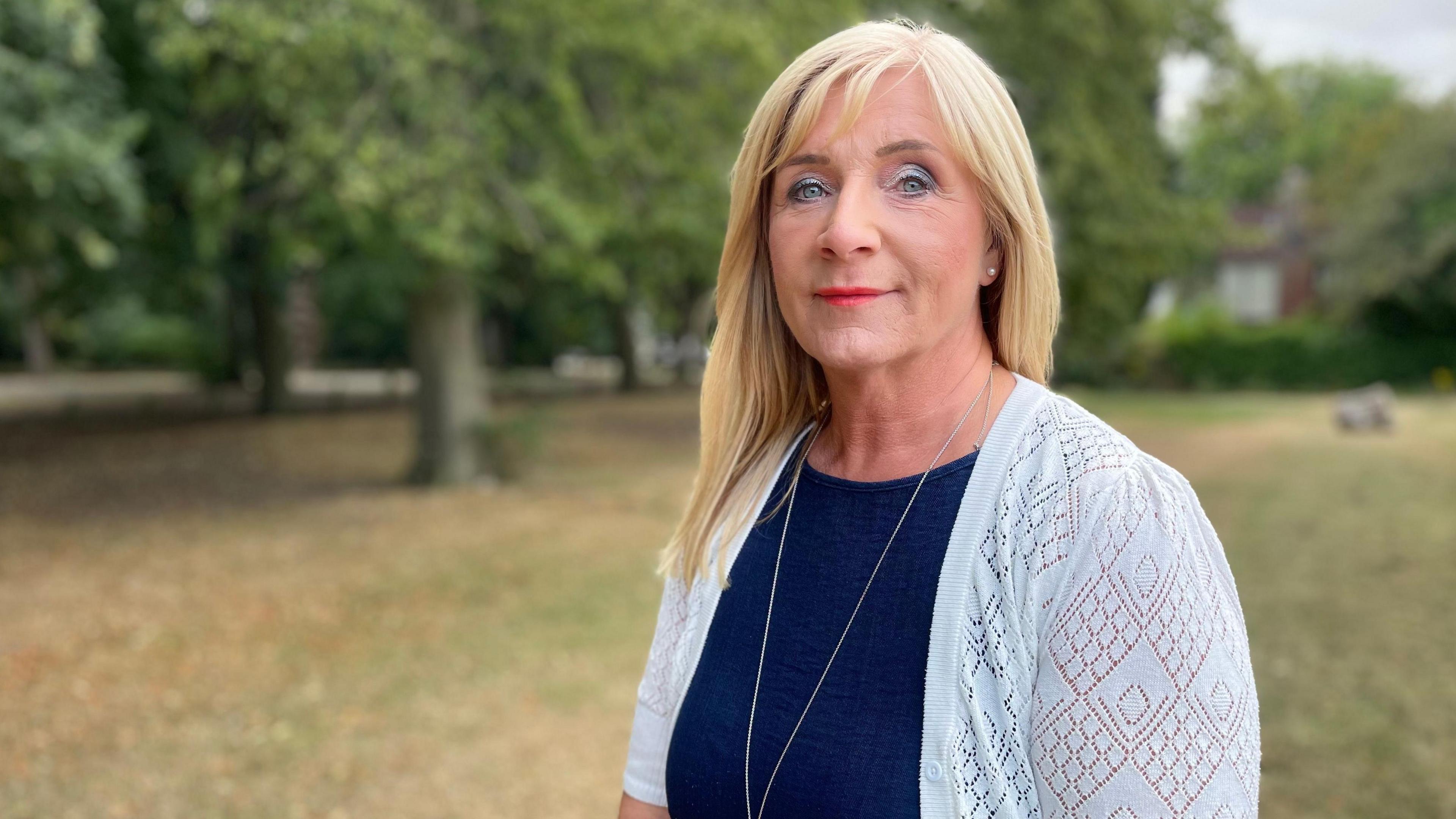 A woman with blond hair and a blue cardigan is standing on a grass field in front of trees 