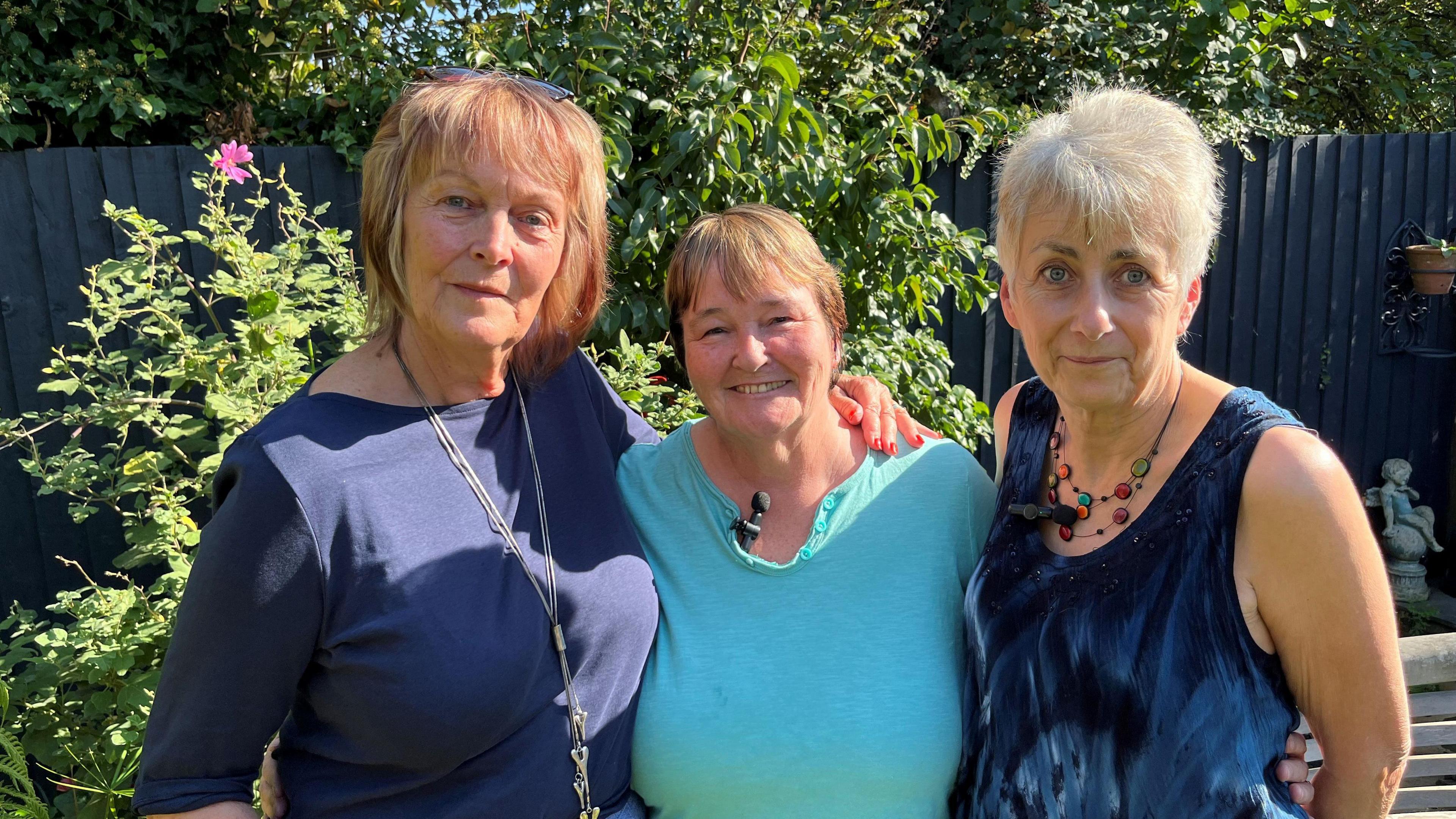 Marie Burgess, Diane Butler and Julia Hatton are all 66, Marie has sandy coloured hair in a bob style, with sunglasses perched on her head, she has a dark blue top on with two silver necklaces hanging down to her midriff. Diane has short auburn hair and is wearing a turquoise round necked top with buttons undone, Julia has short light brown hair with a sleeveless blue dress on and multi-coloured beads around her neck, behind the ladies is a garden fence and various shrubs and trees.