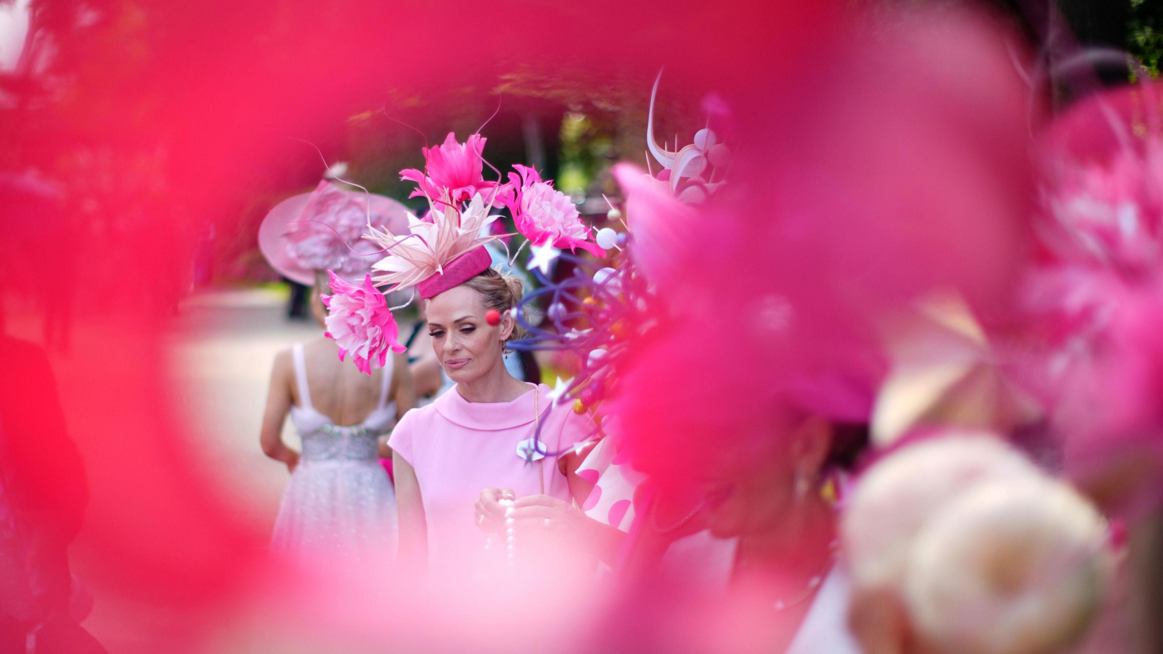 A woman, who is wearing a pink hat and a pink dress, is seen through a woman's pink fascinator.