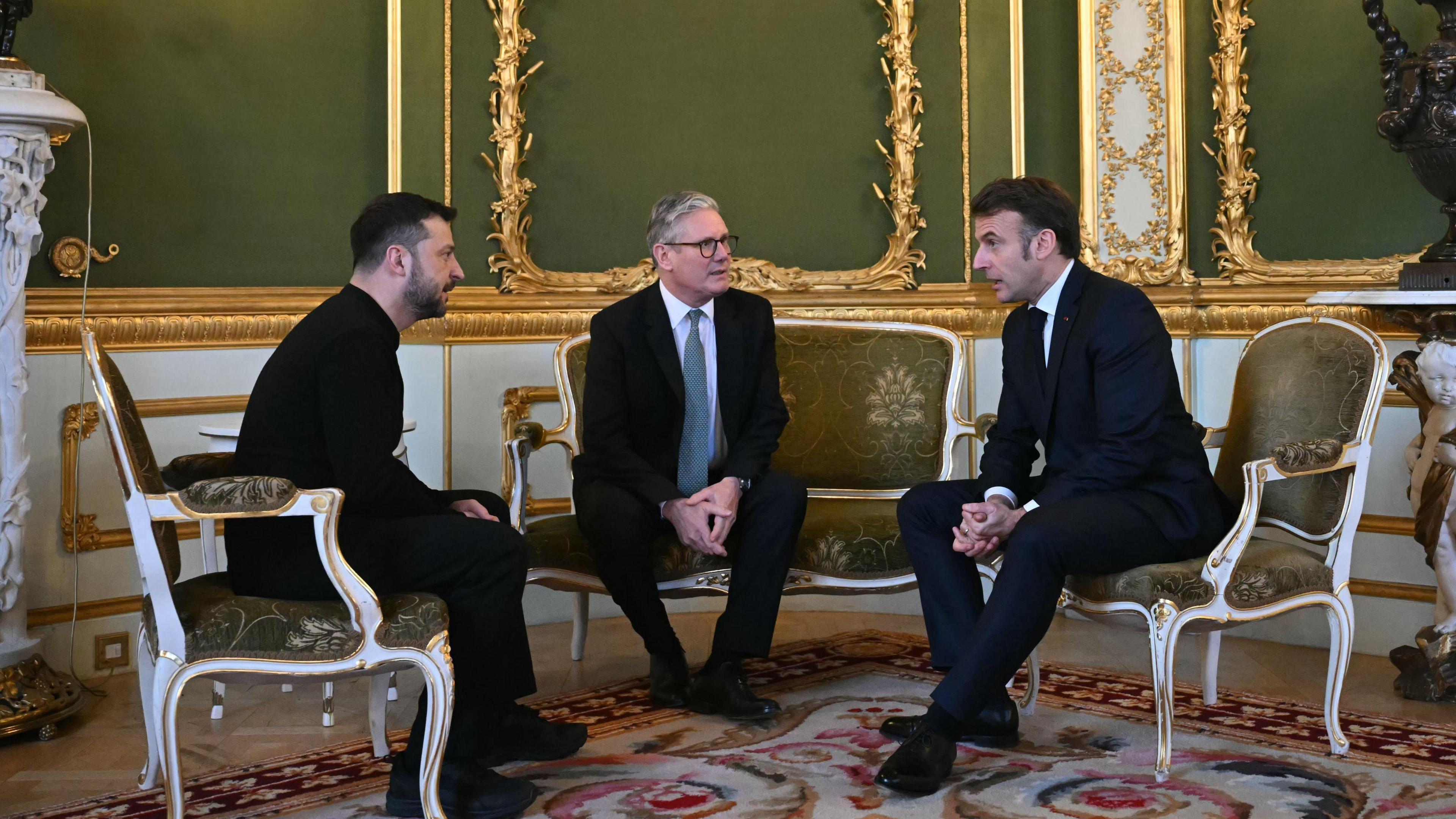 Volodymyr Zelensky, Sir Keir Starmer and Emmanuel Macron sitting on white and green chairs in a white and green panelled room with gold detail.