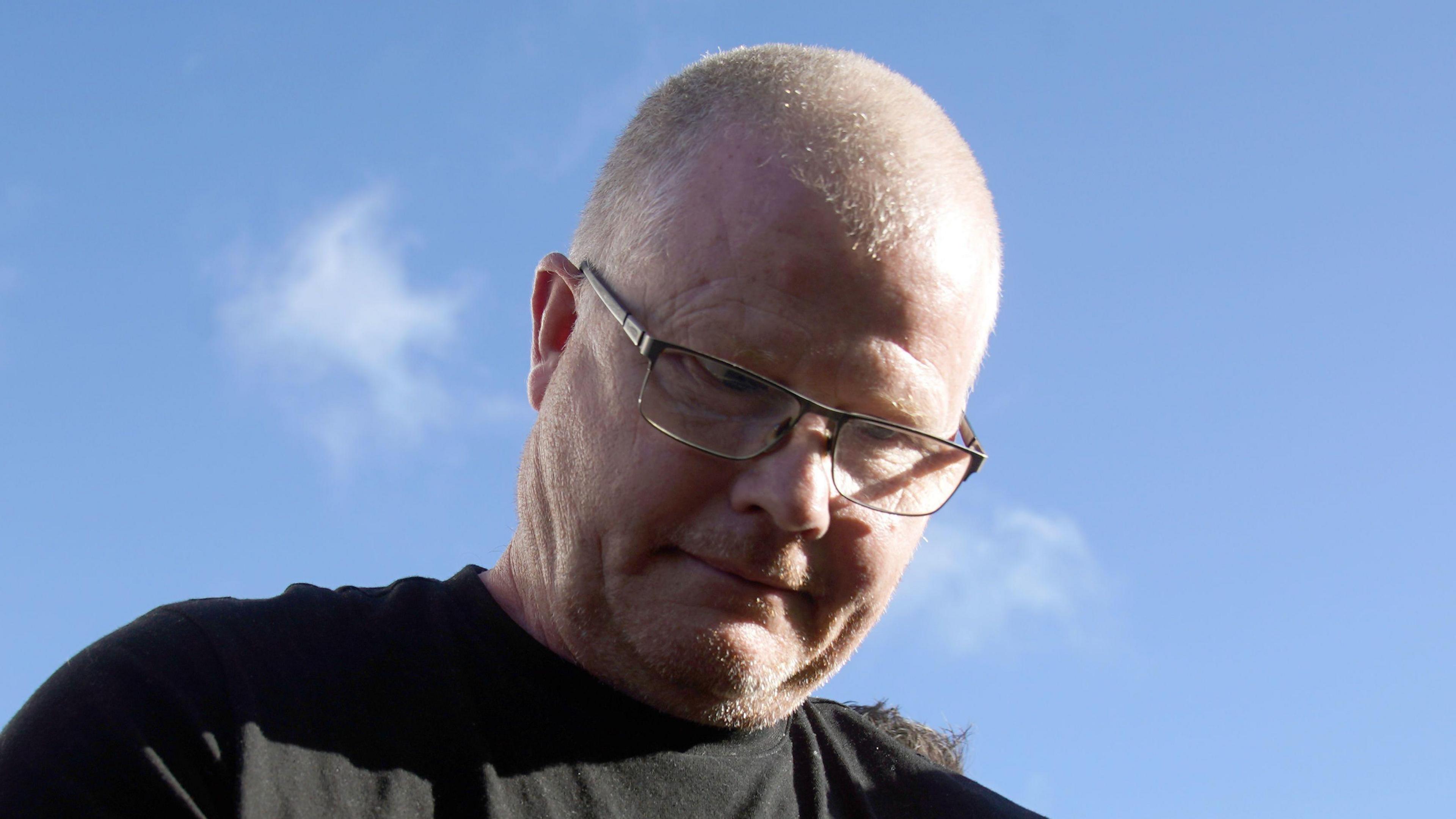 Richard Satchwell is wearing a black t-shirt and he has a bald head with square glasses on. He is looking down. The sky behind him is bright blue.