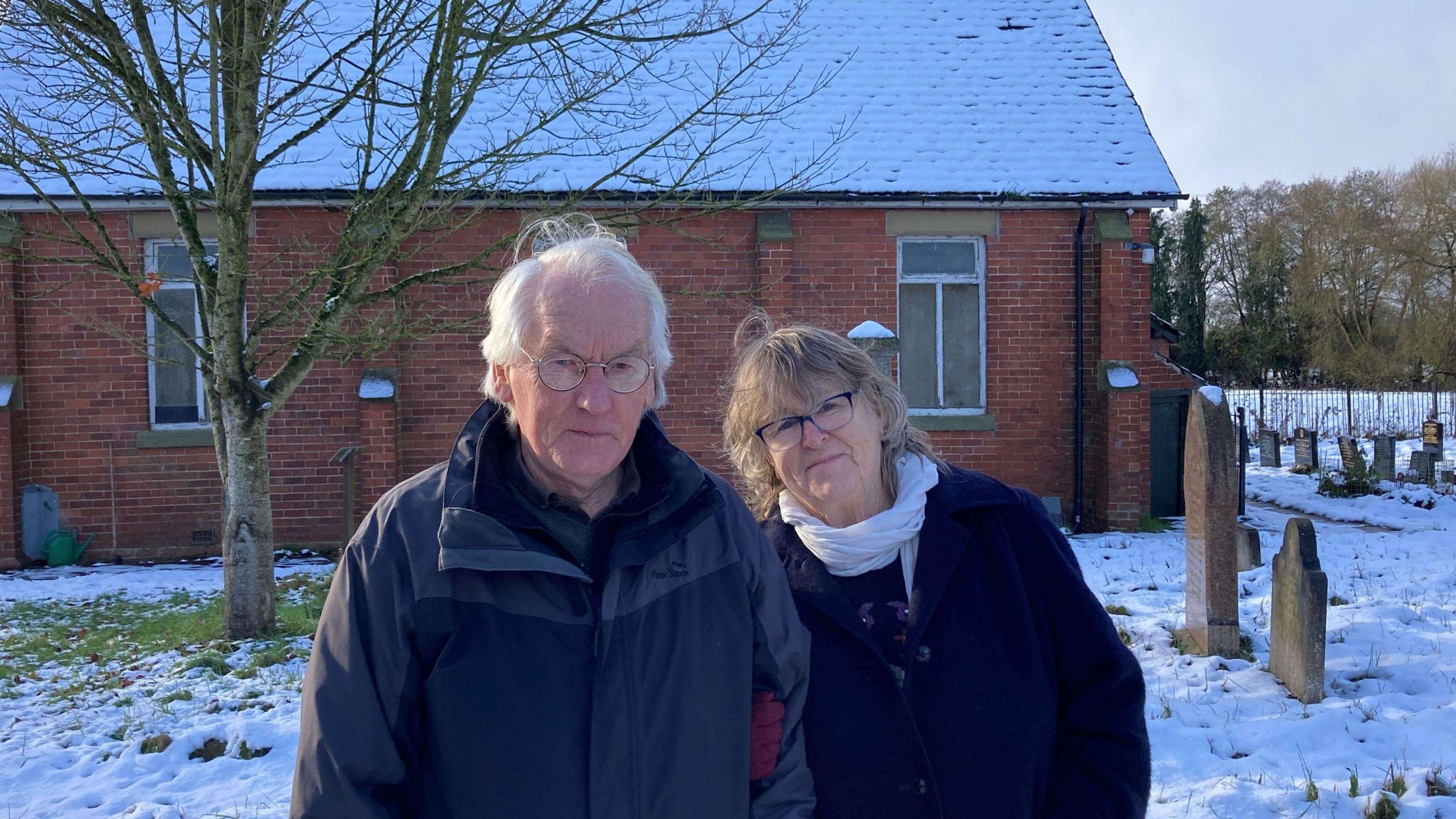 Couple standing in front of a disused chapel in a snowy graveyard, surrounded by headstones.