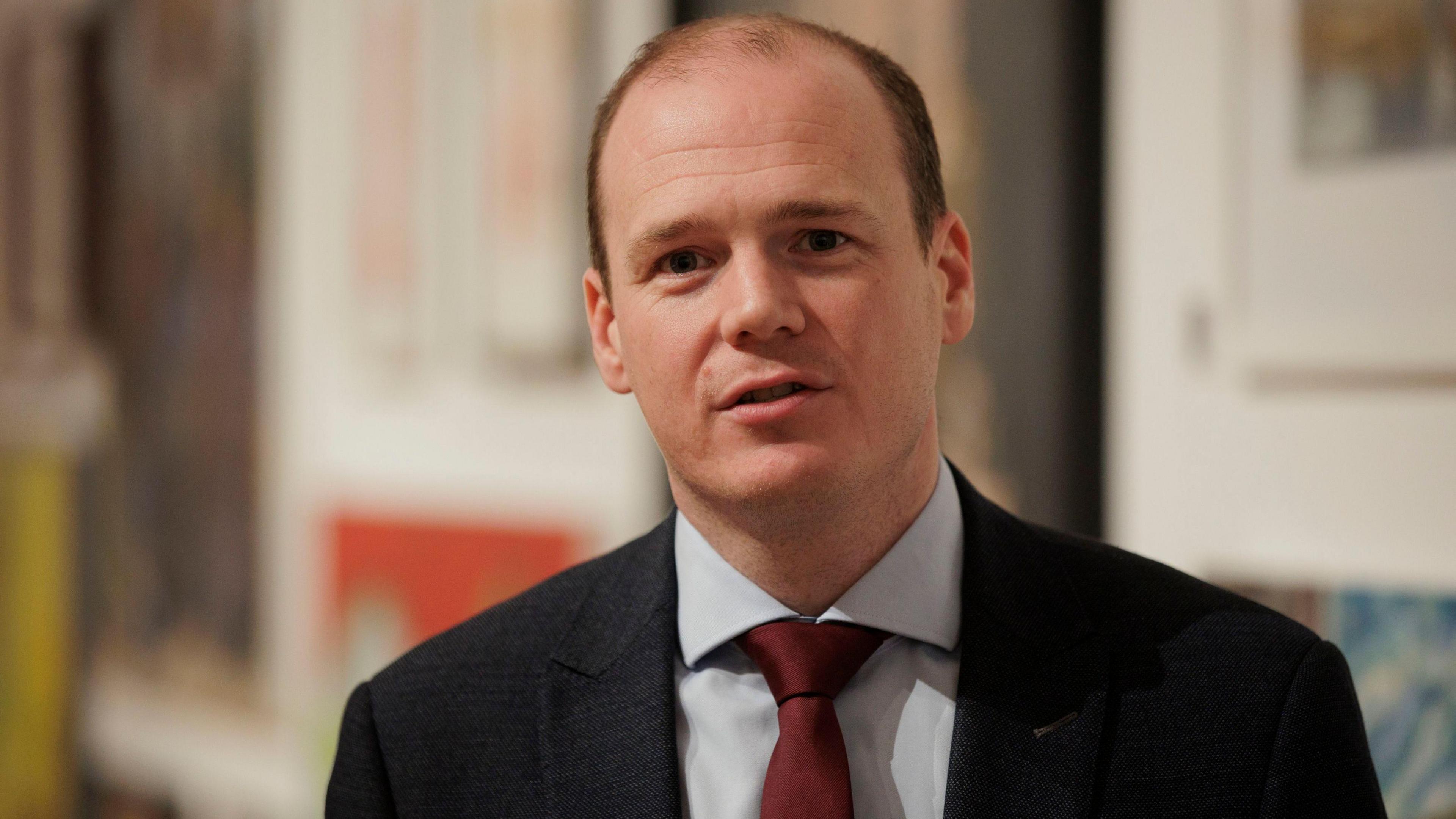 Gordon Lyons wears a black suit, grey shirt and red tie. He has brown hair.