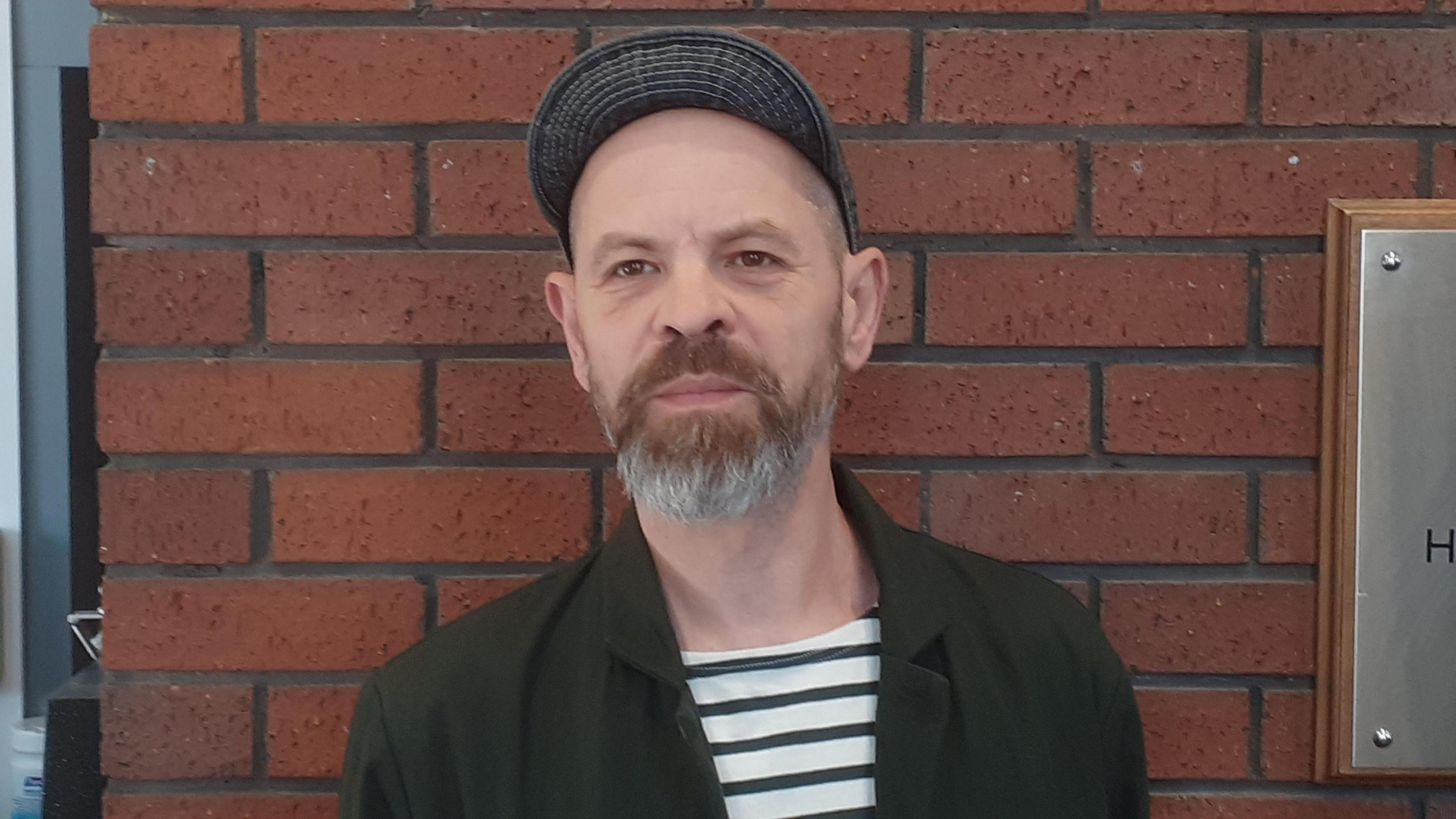 A man in a black and white striped top and black overcoat stands by a brick wall