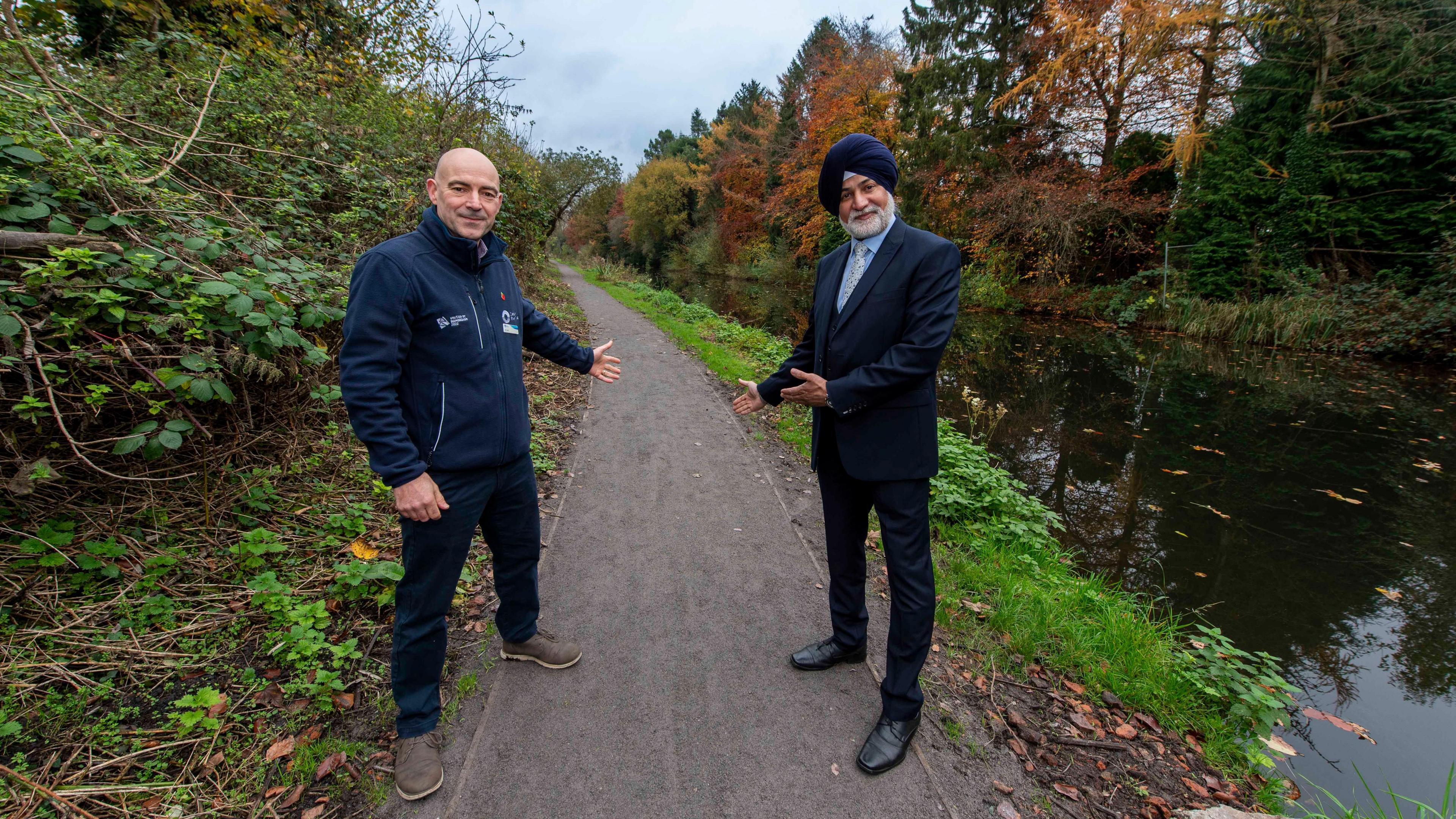 Two men stand on a newly-resurfaced path by the side of a canal. One man is wearing a suit and a turban, the other is wearing a navy blue jacket and navy blue trousers and brown shoes.