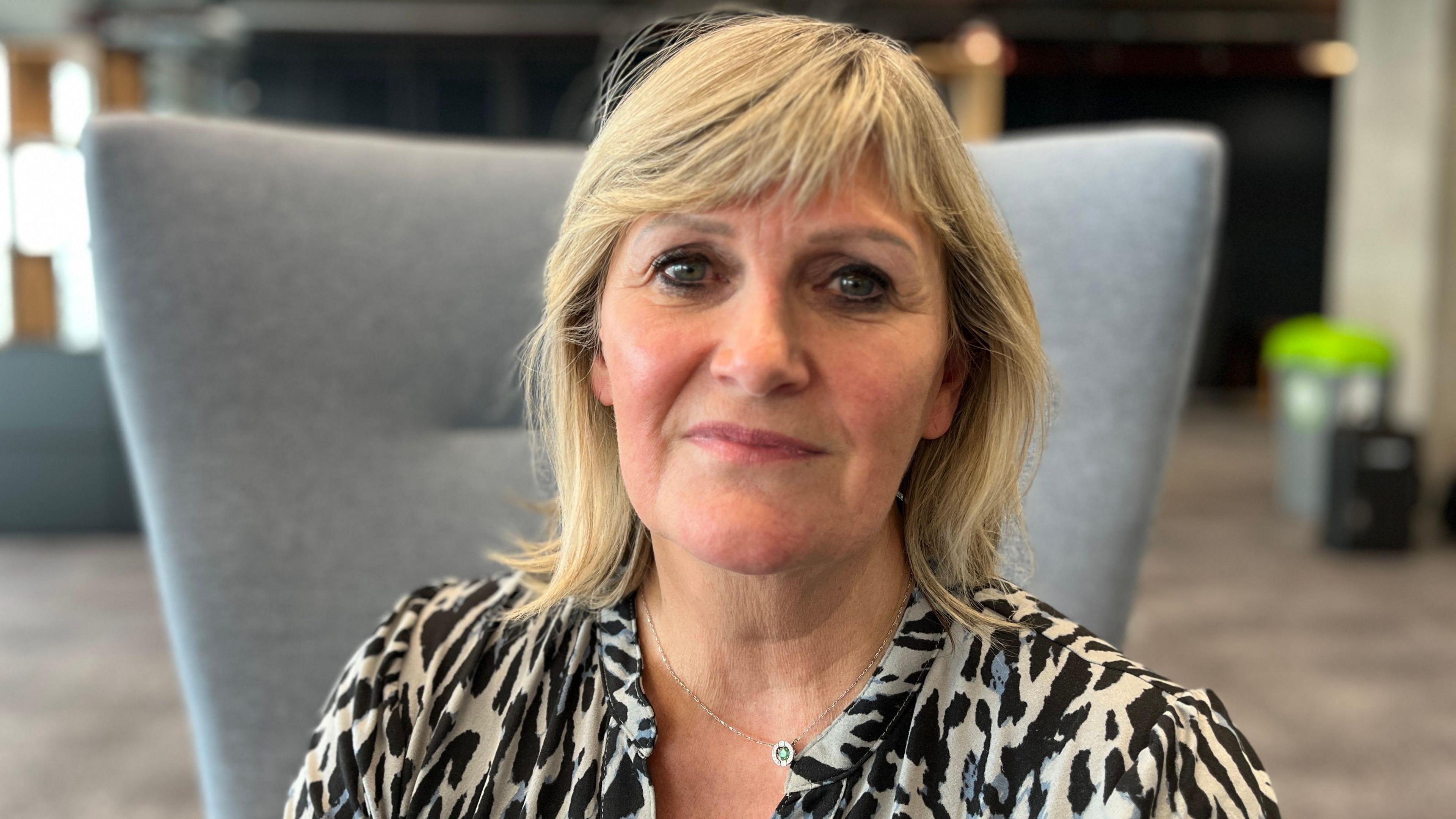 A woman with blonde hair, wearing a black-and-white top and a necklace, sits on a grey chair in an office