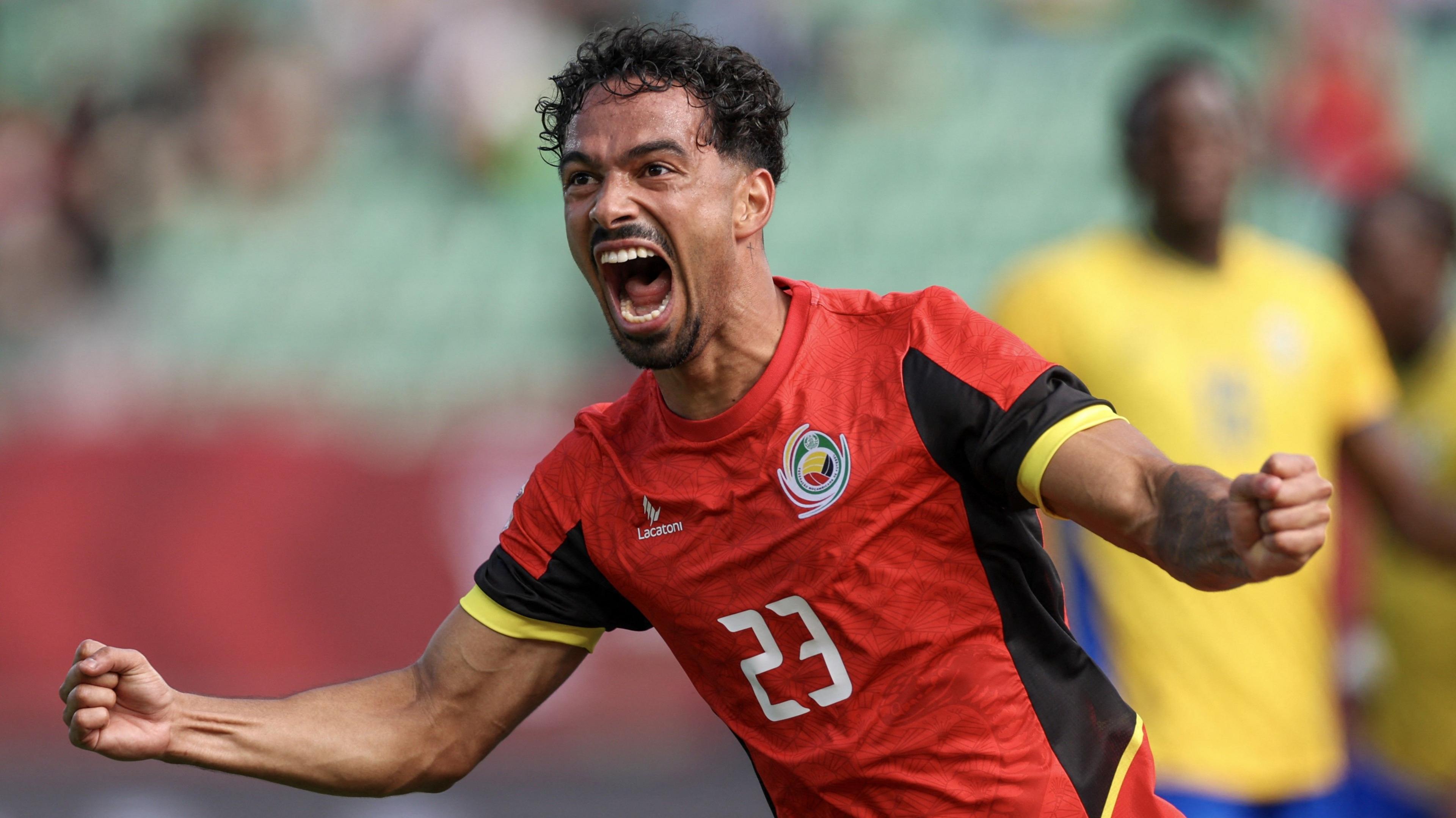 Diogo Calila shown from waist up wheeling away in celebration after scoring a goal for Mozambique against Gabon at Afcon 2025. He is wearing a short-sleeved red jersey with black and yellow trim and a white number 23 on the centre of the chest. Calila has a thin goatee beard and moustache and is opening his mouth wide to scream in joy, while spreading both arms out to the side and clenching the fists. Gabon players in yellow and blue kit are blurred in the background