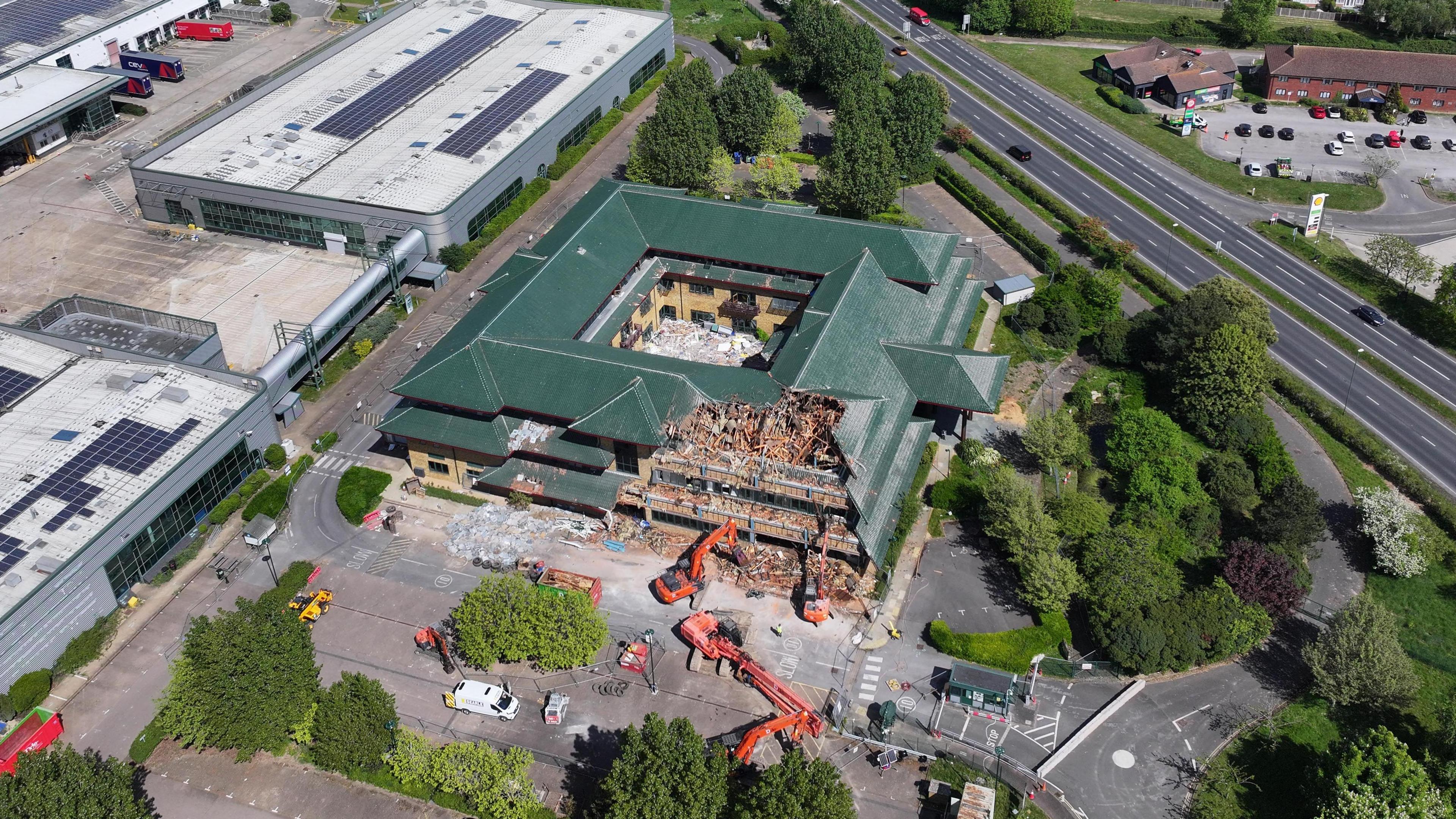 An aerial image of demolition works on a square building with a dark green roof. The building is surrounded by cranes and has a chunk taken out of its side. the roof has a number of triangular layers.