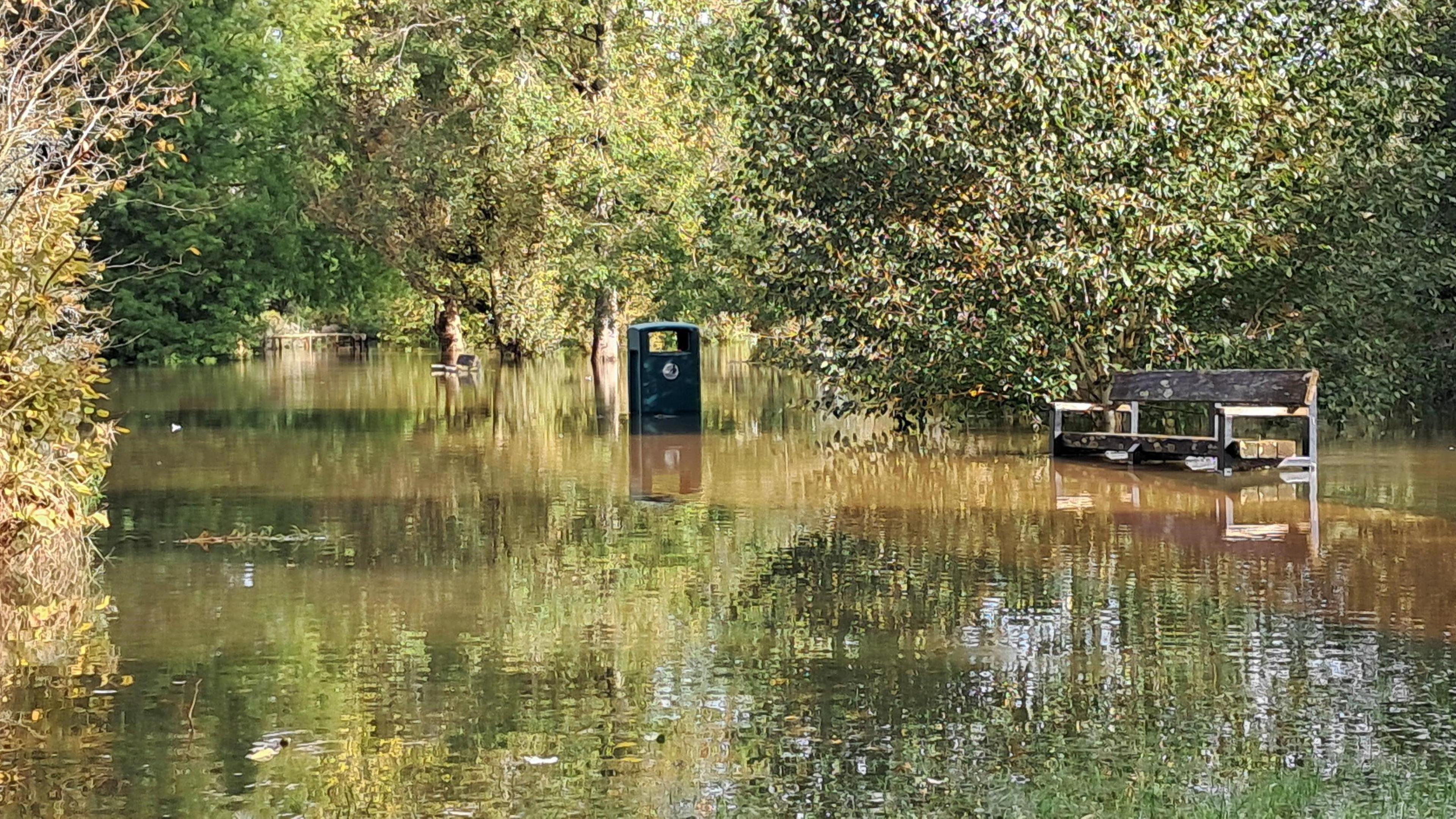 What looks like a path which has been flooded. The water level is several inches deep and has risen to the height of the seat on a wooden bench at the side of the bath. A bin is also sitting in the middle of the floodwater.