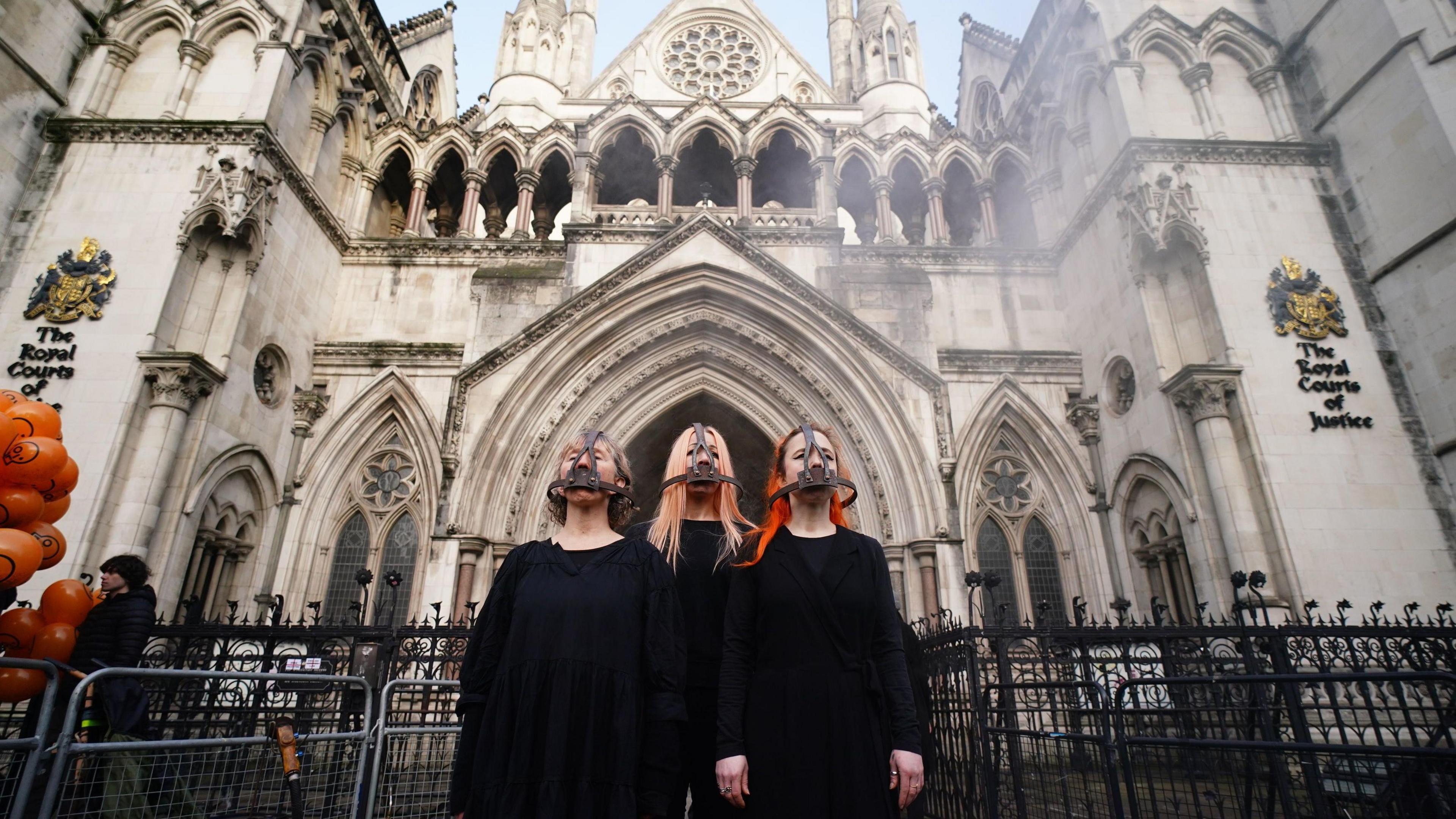 Three campaigners stand in black outside court, wearing metal head-gear which covers their mouths 