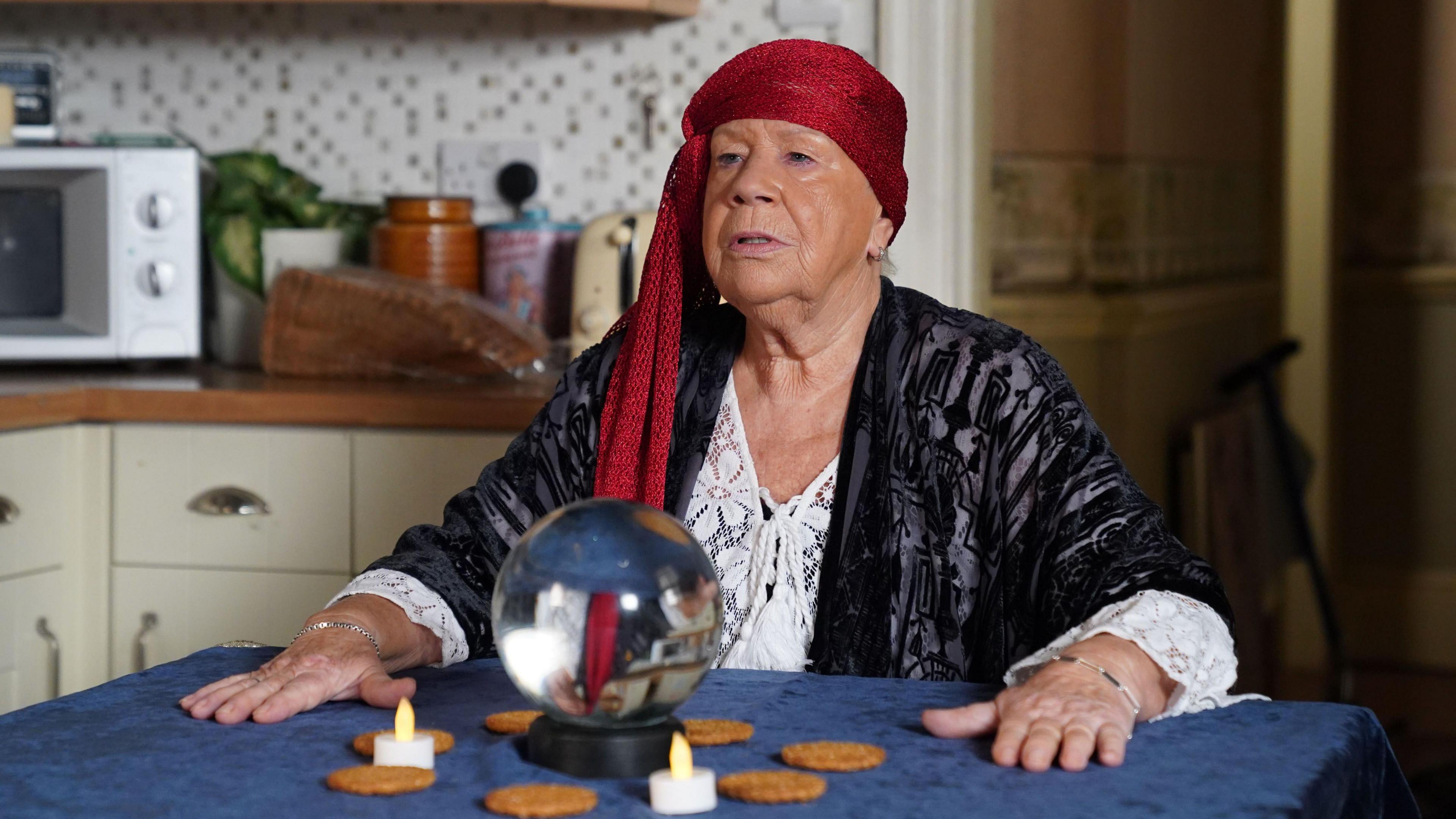 Laila Morse is wearing a red headband. She is sat at a table with a crystal ball and her hands are placed wither side of it. The crystal ball is surrounded by a circle of tea lights and biscuits.