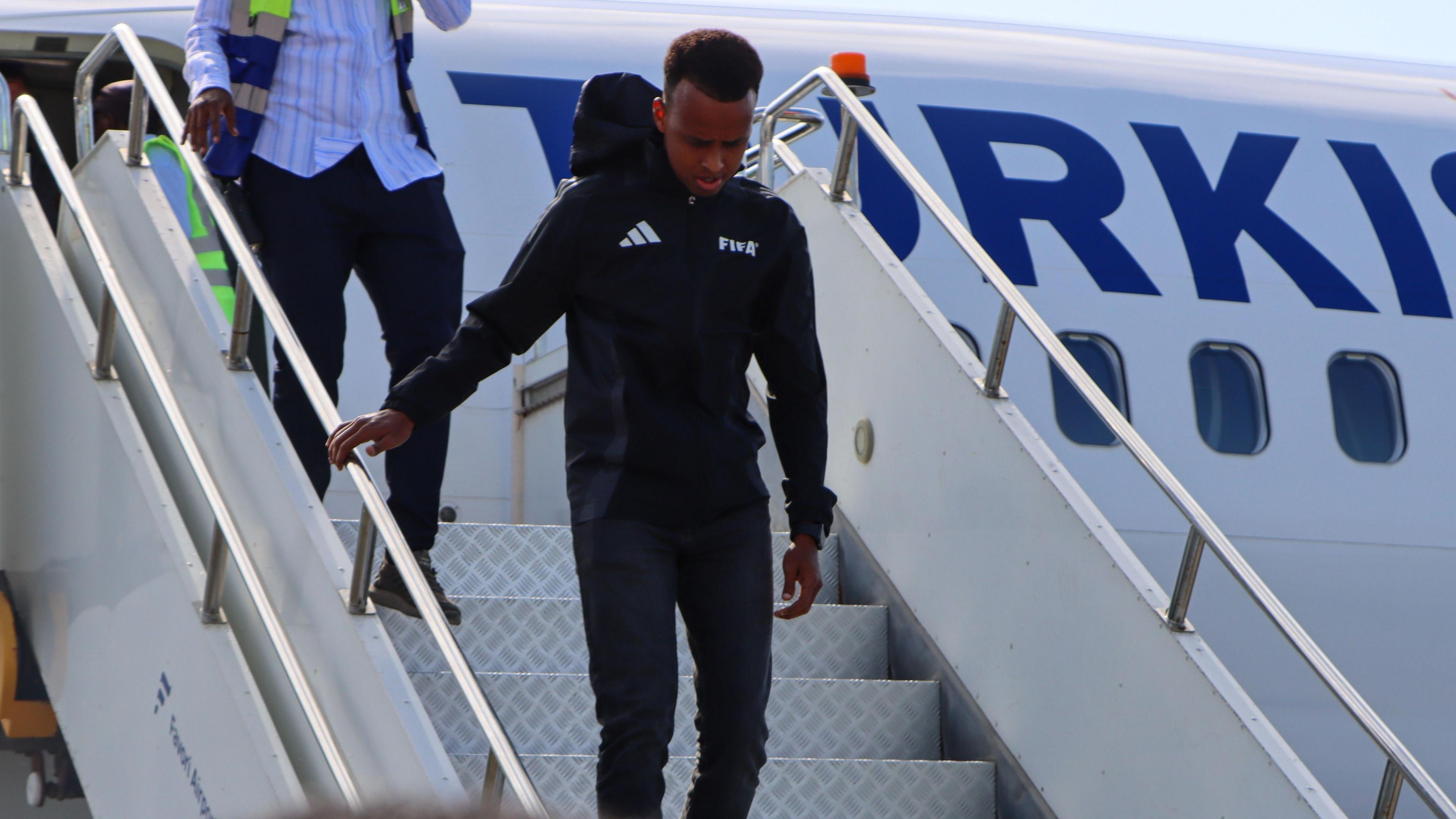 Omar Artan, wearing a black Fifa hoodie and trousers, walks down the white metal stairs from the white Turkish Airlines plane