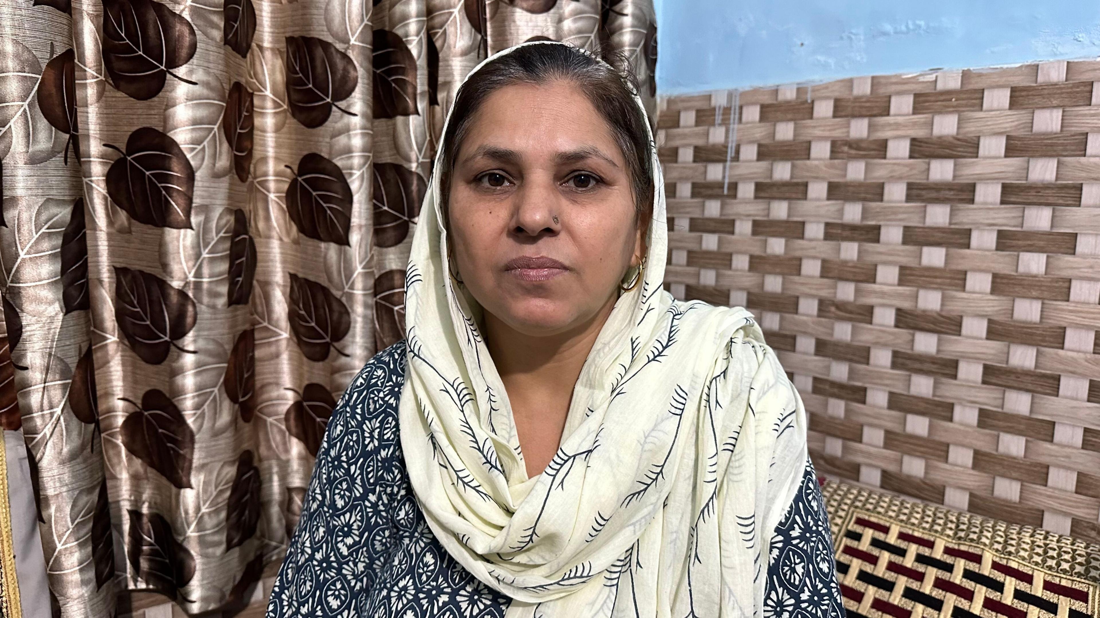 Harshita Brella's mother Sudesh Kumari looks into the camera. She is wearing a white printed scarf on her head and a blue kurta.