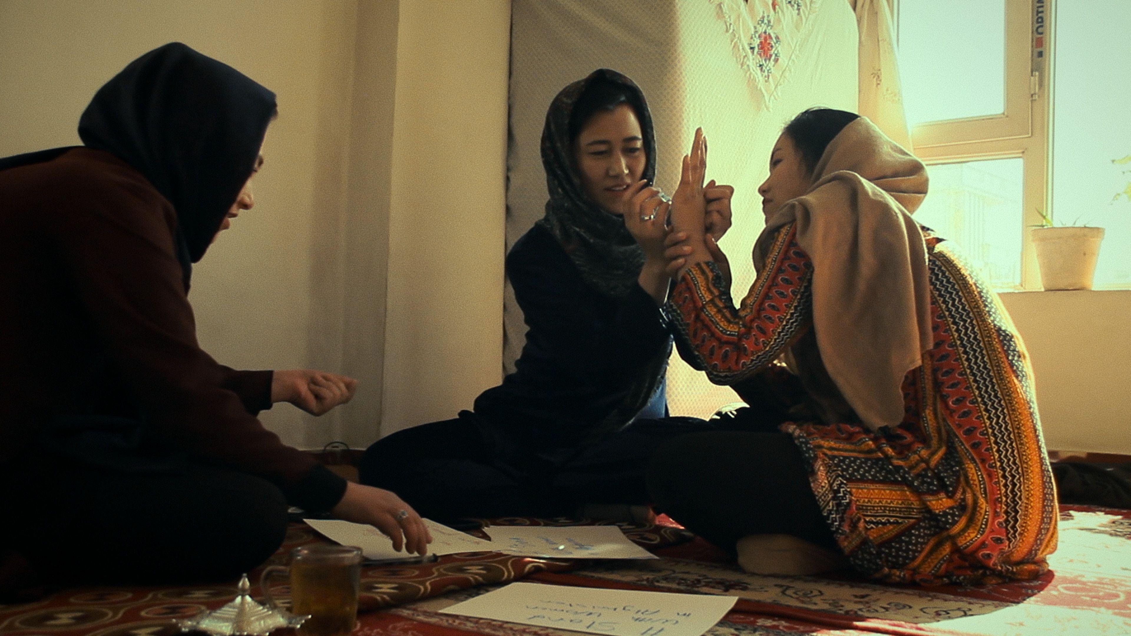 A still from Bread & Roses showing three women sitting together on the floor. The woman in the middle writes on the hand of the woman sitting to her left while the third woman looks at sheets of paper spread on the carpet. 