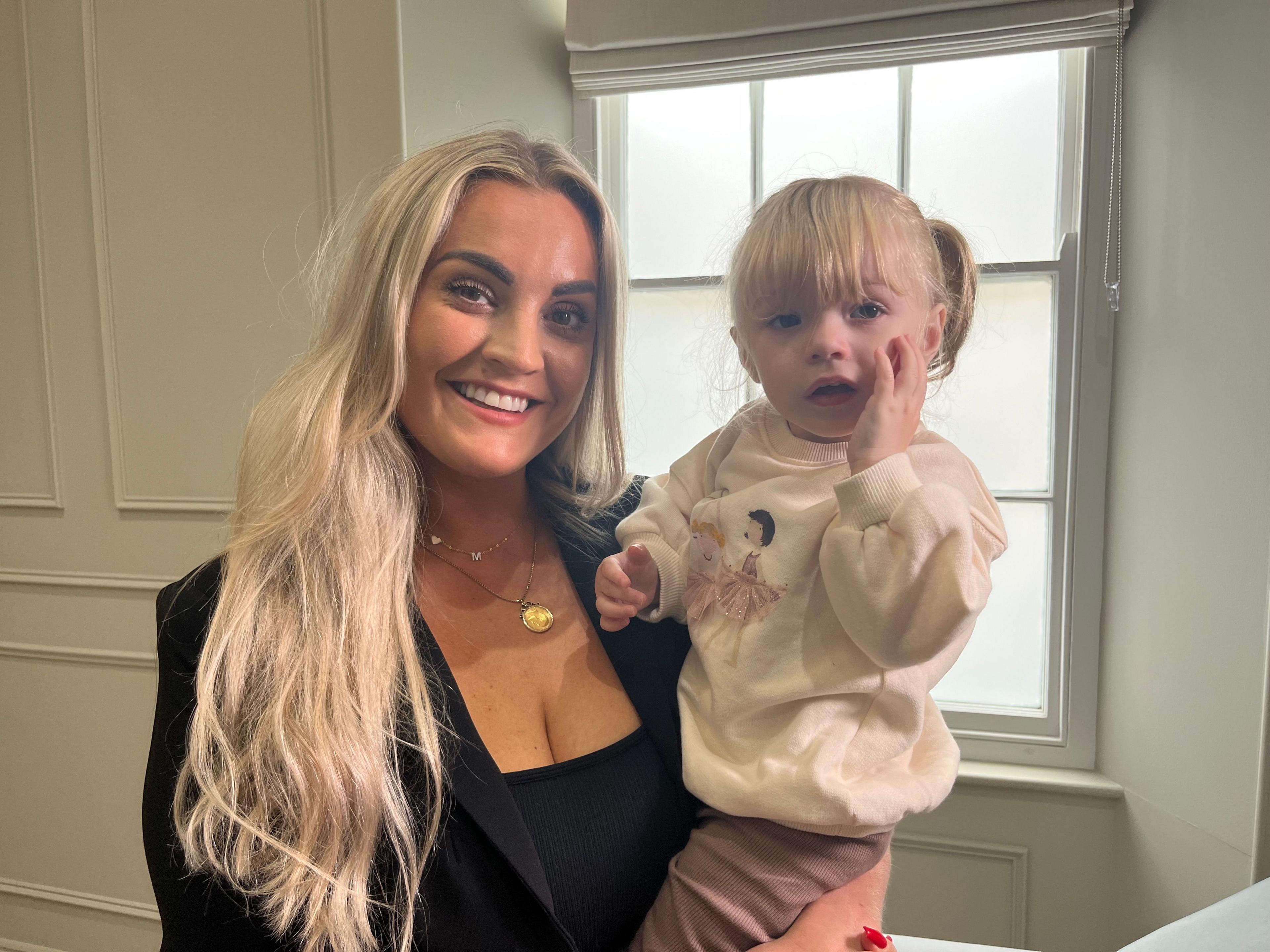 Sophie smiling, holding her daughter Mimi as she stands in a room, in front of a window