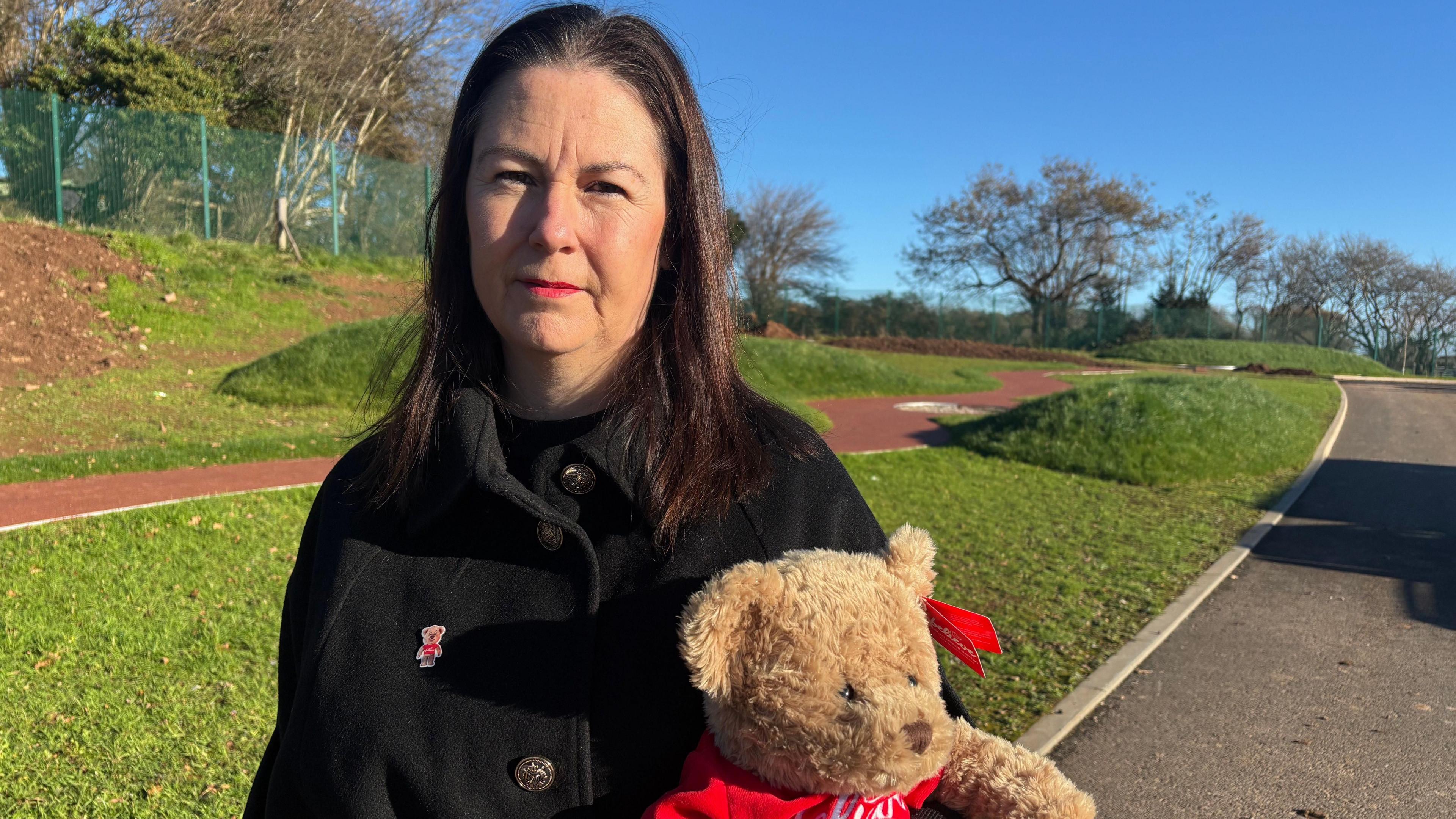 A woman with long dark hair stands in a garden holding a teddy bear. She is wearing a black coat with a bear symbol pinned to it.