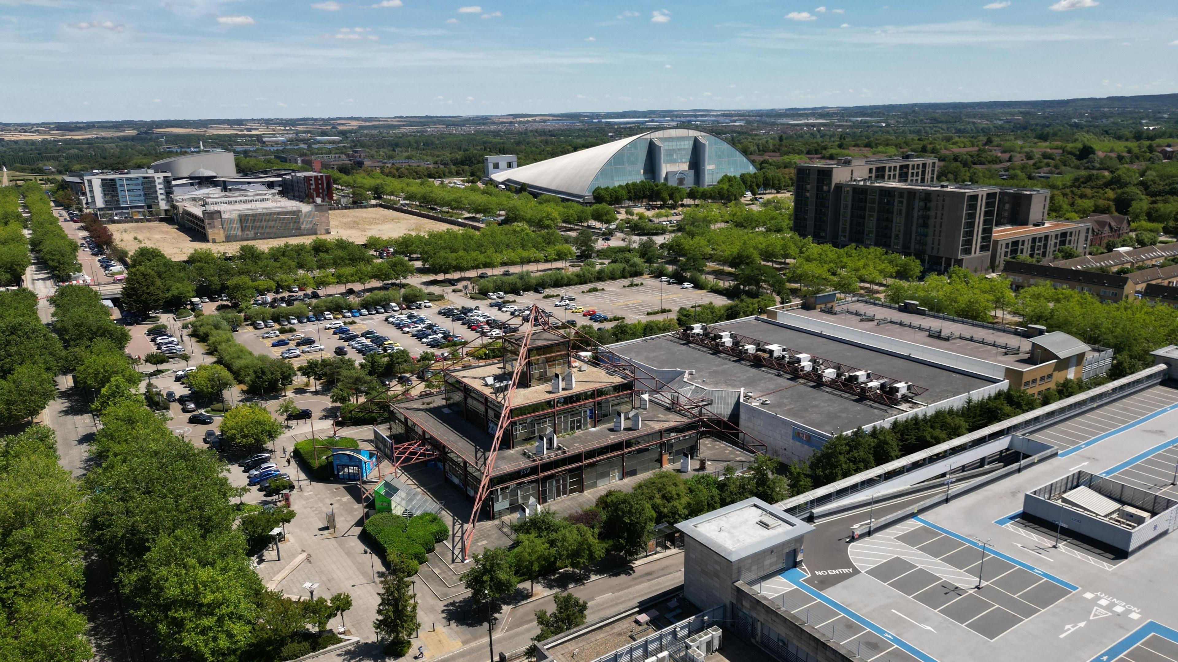An aerial drone view of Milton Keynes with a dome shaped building in the distance. There are large buildings and a surface car park as well as trees.