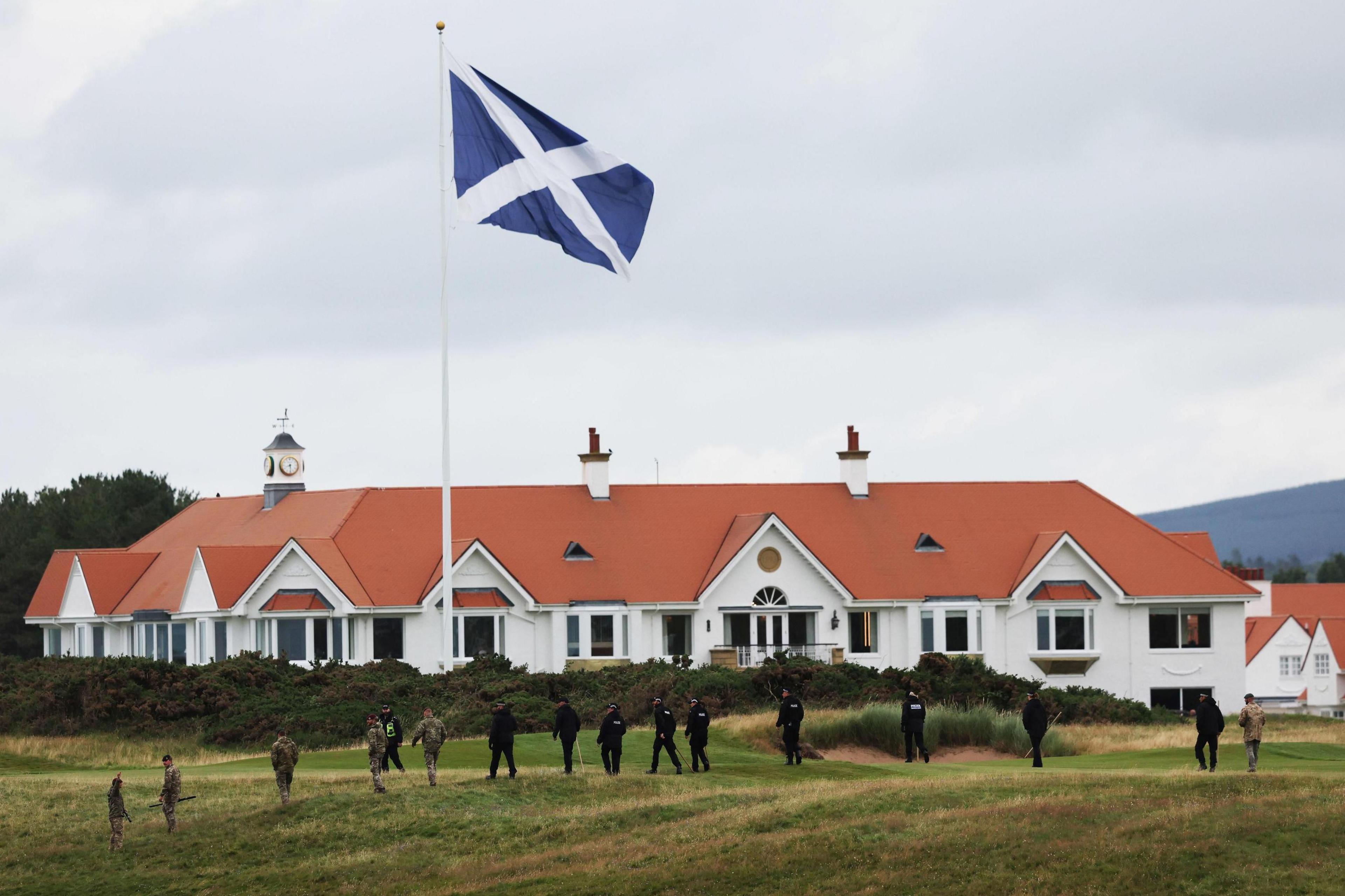 Police officers and military personnel search the area at the Turnberry golf course in South Ayrshire. They are walking in front of a large Saltire on a flag pole and a building with white walls and a terracotta coloured roof.