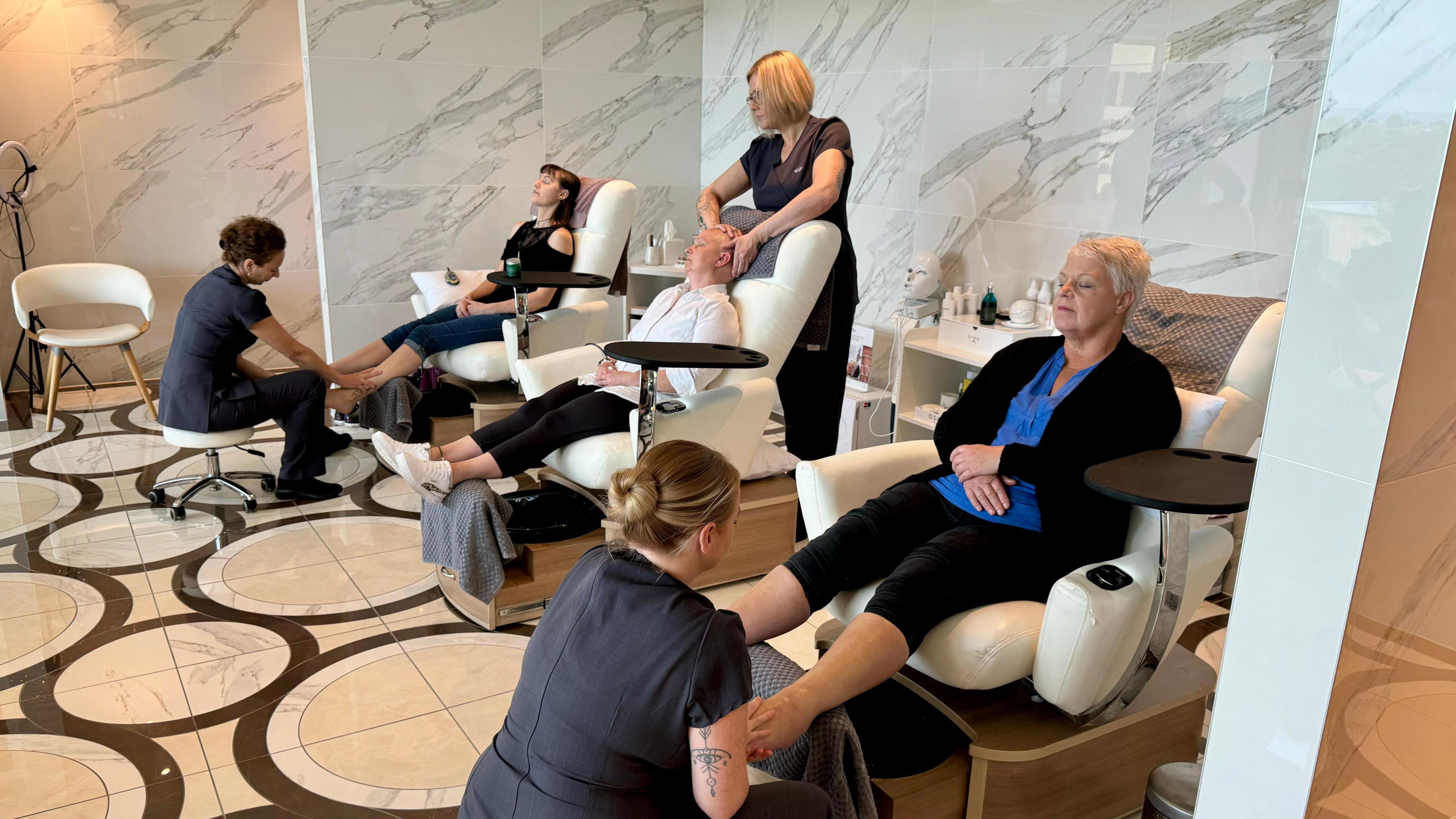 Three women sit on chairs in a spa with an ornate marble floor having their feet massaged by staff