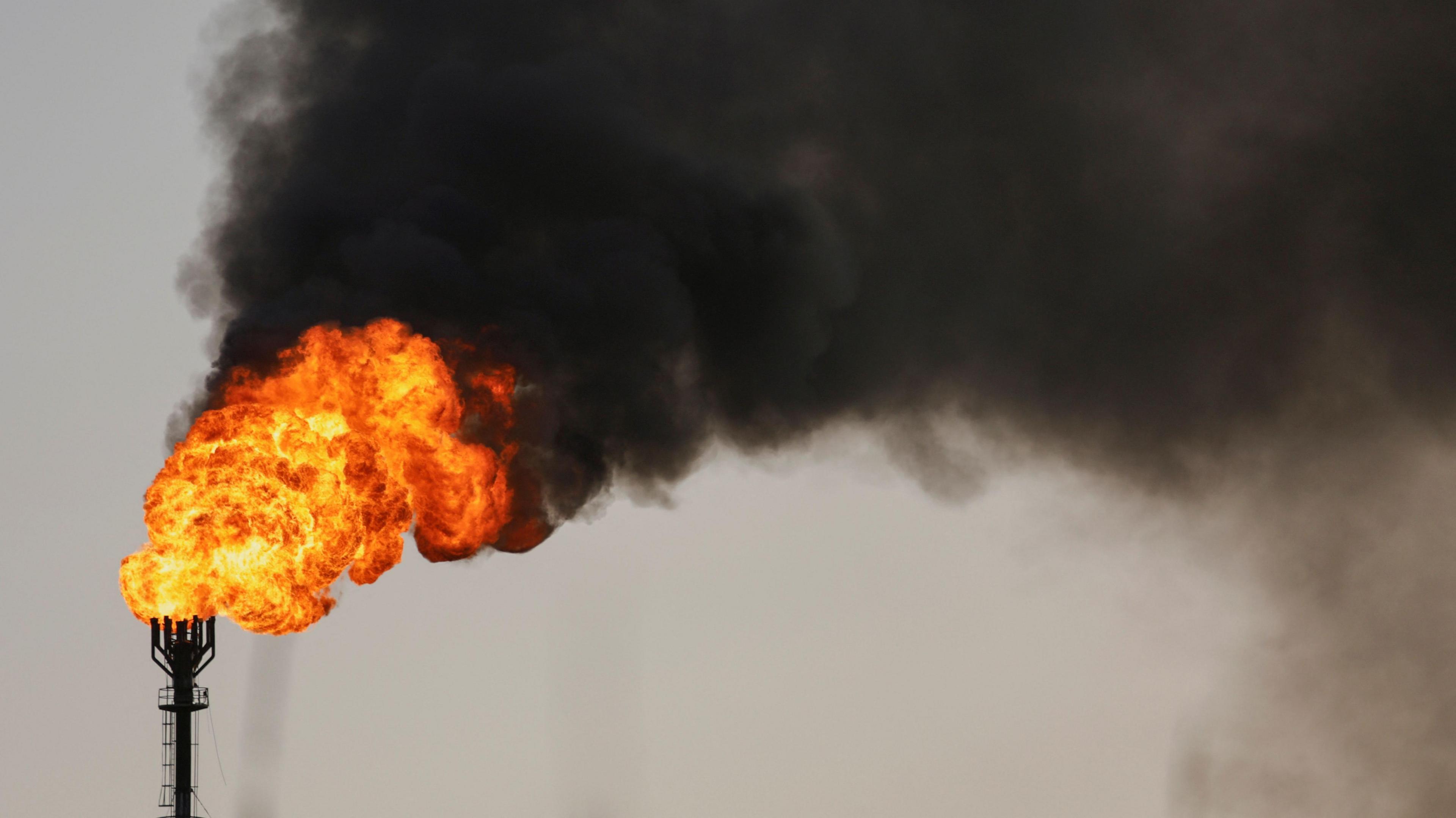 Blooms of bright orange fire leading to thick black smoke burst out from a metallic pipe in a grey sky.