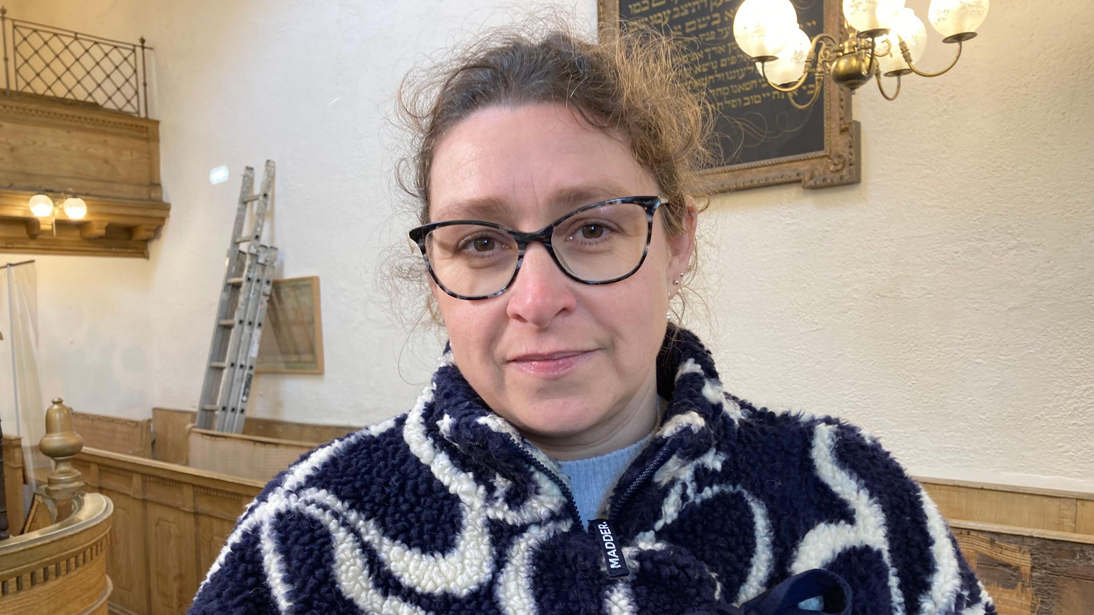 Abigail Fisher pictured inside a temple. She has curly hair tied back and wears large dark-framed glasses. She is wearing a navy and white fluffy fleece with a distinctive swirling pattern on it. 