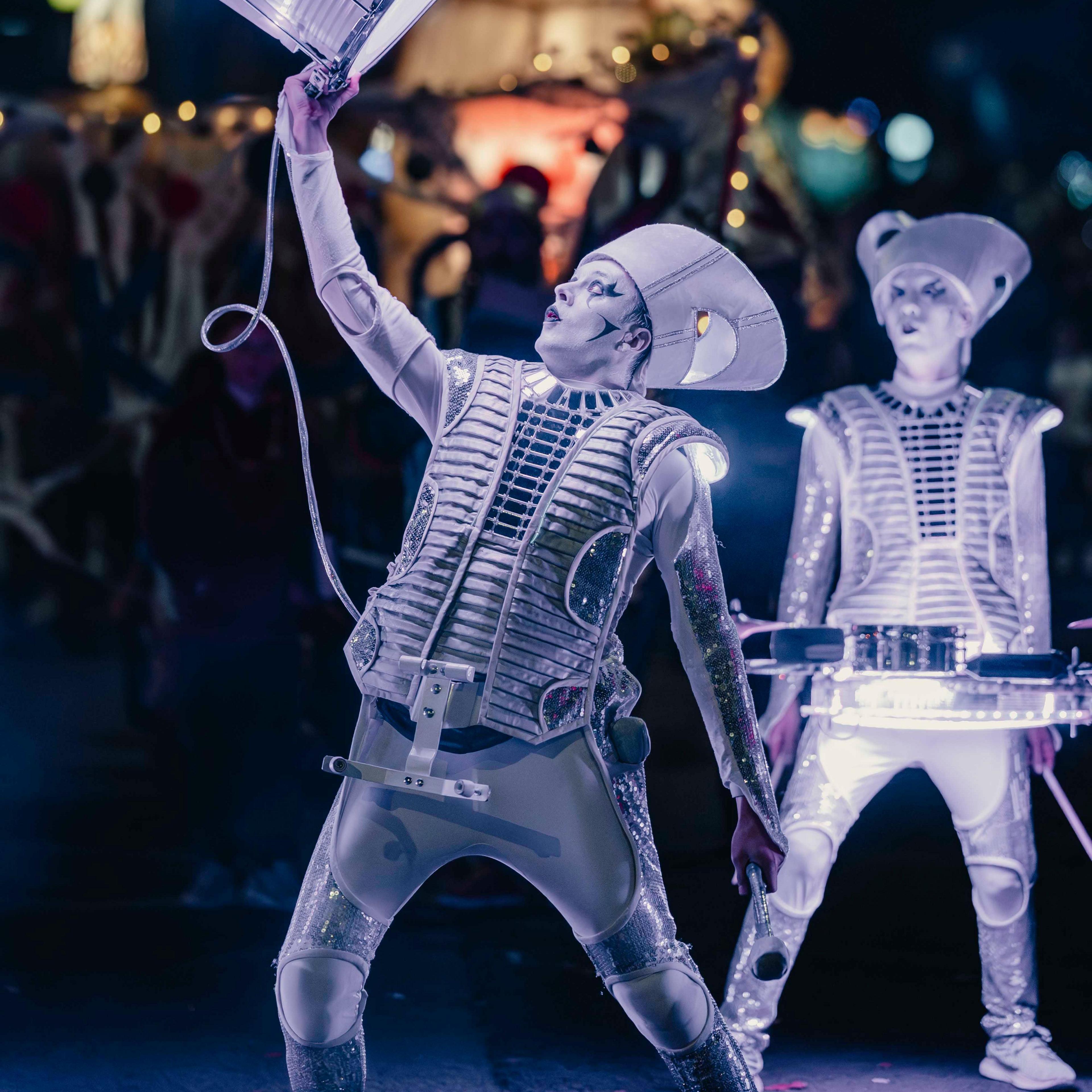 Two drummers dressed in white military like uniforms and cone-shaped hats perform against a black night sky. Their drums are wrapped in bright white LED lights.