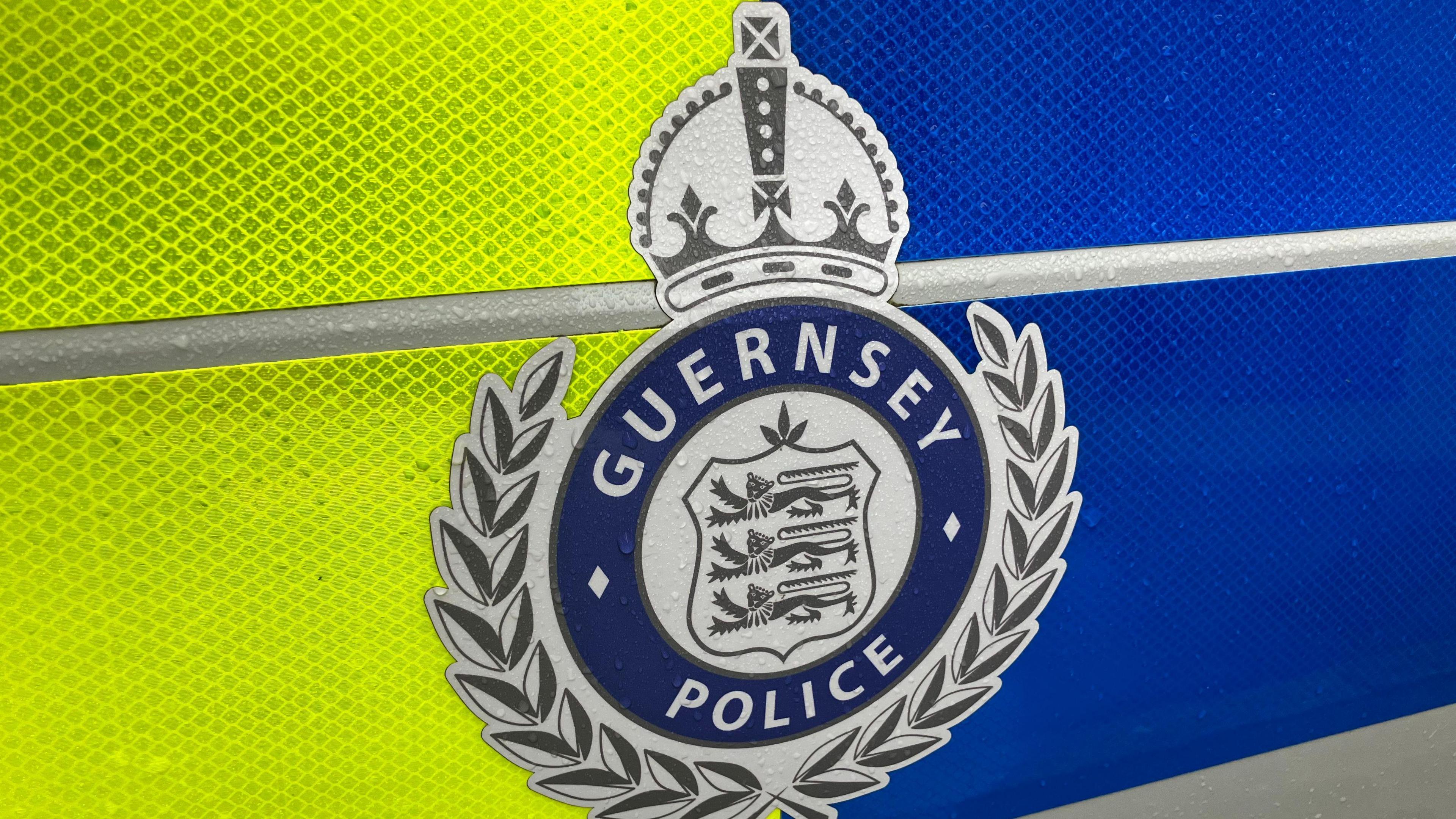 The Guernsey Police logo of three lions on a coat of arms, surrounded by a laurel wreath and topped by a crown, with the word Guernsey Police around it is posted over a fluorescent green and blue background in a close up of the side of a police car.