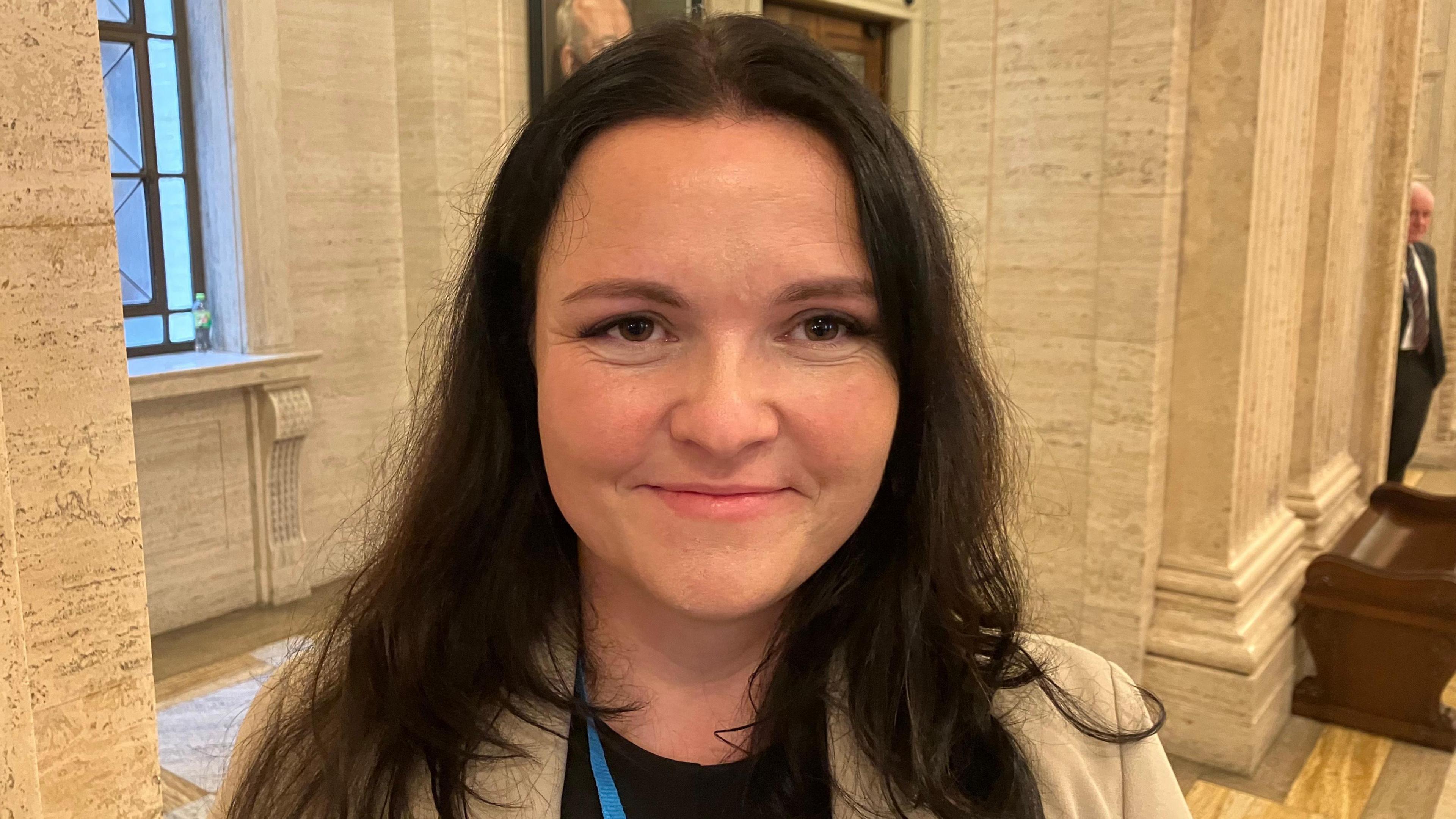 Christine Mhic Colaim is standing in Stormont's Great Hall in front of white, marble pillars. She is a young woman who is smiling at the camera. She has long, brown hair and is wearing a light coloured blazer and a black t-shirt.